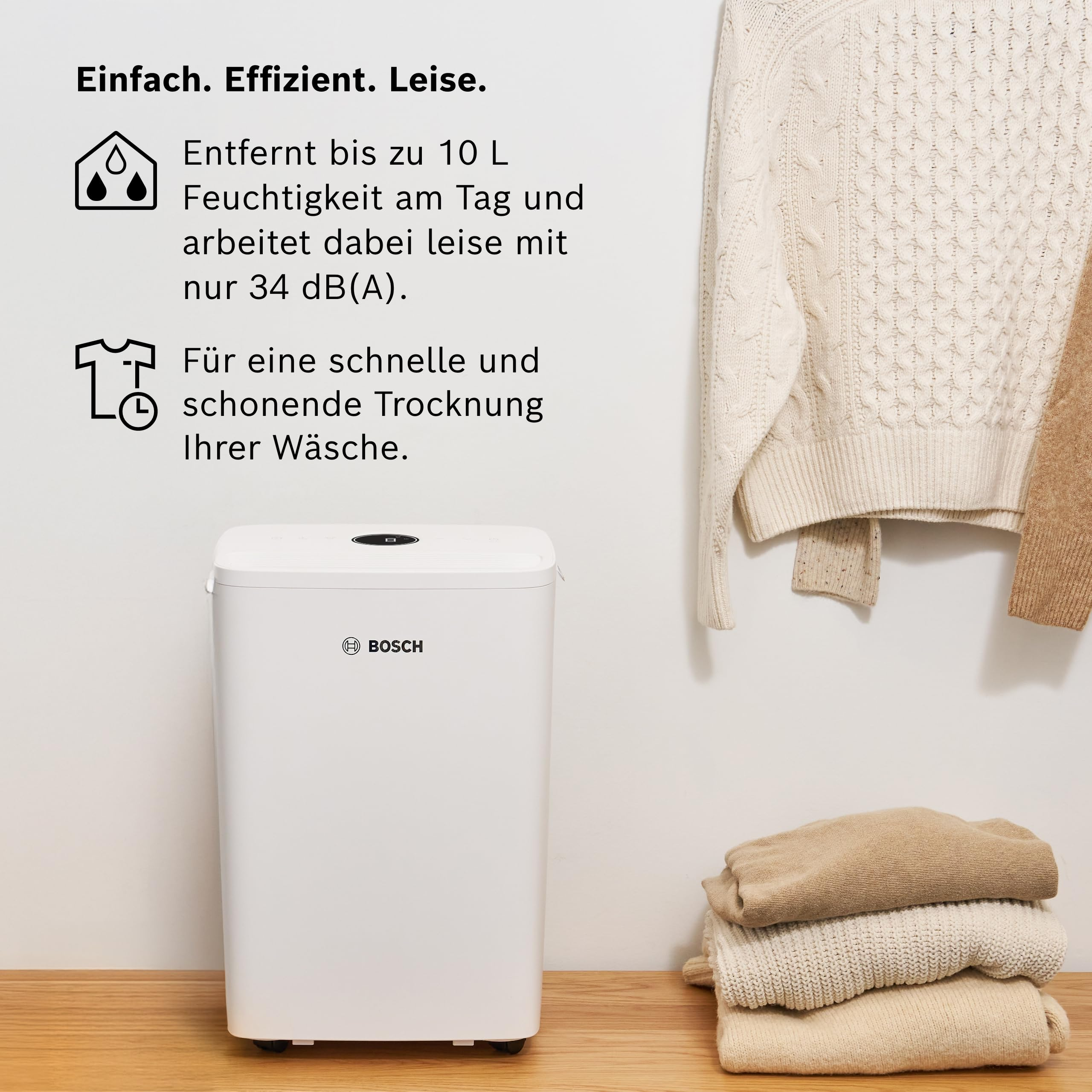 Weißer Bosch Luftentfeuchter auf Holzboden mit gestapelten und hängenden Pullovern.