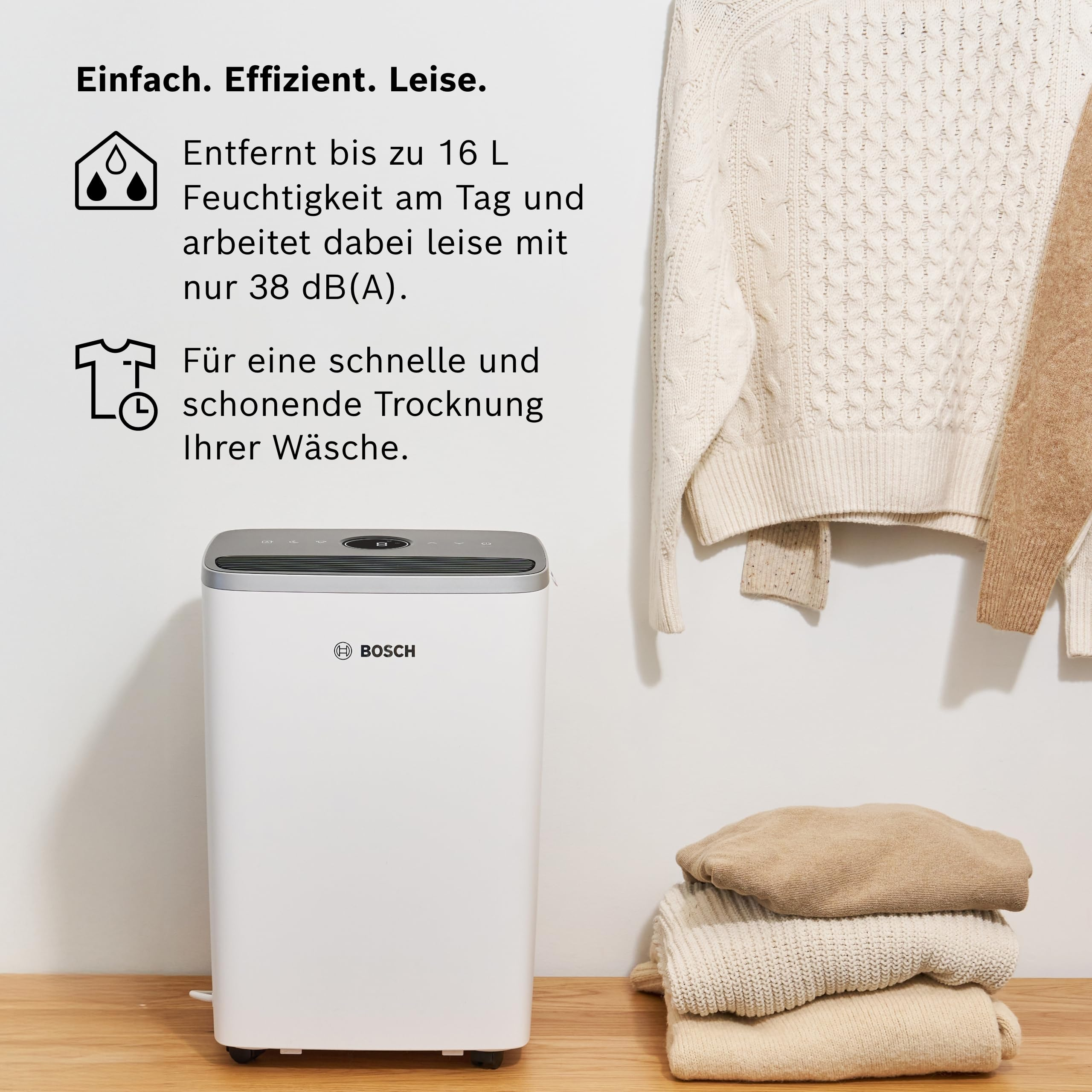 Ein weißer Luftentfeuchter von Bosch steht neben gestapelter und hängender Kleidung.