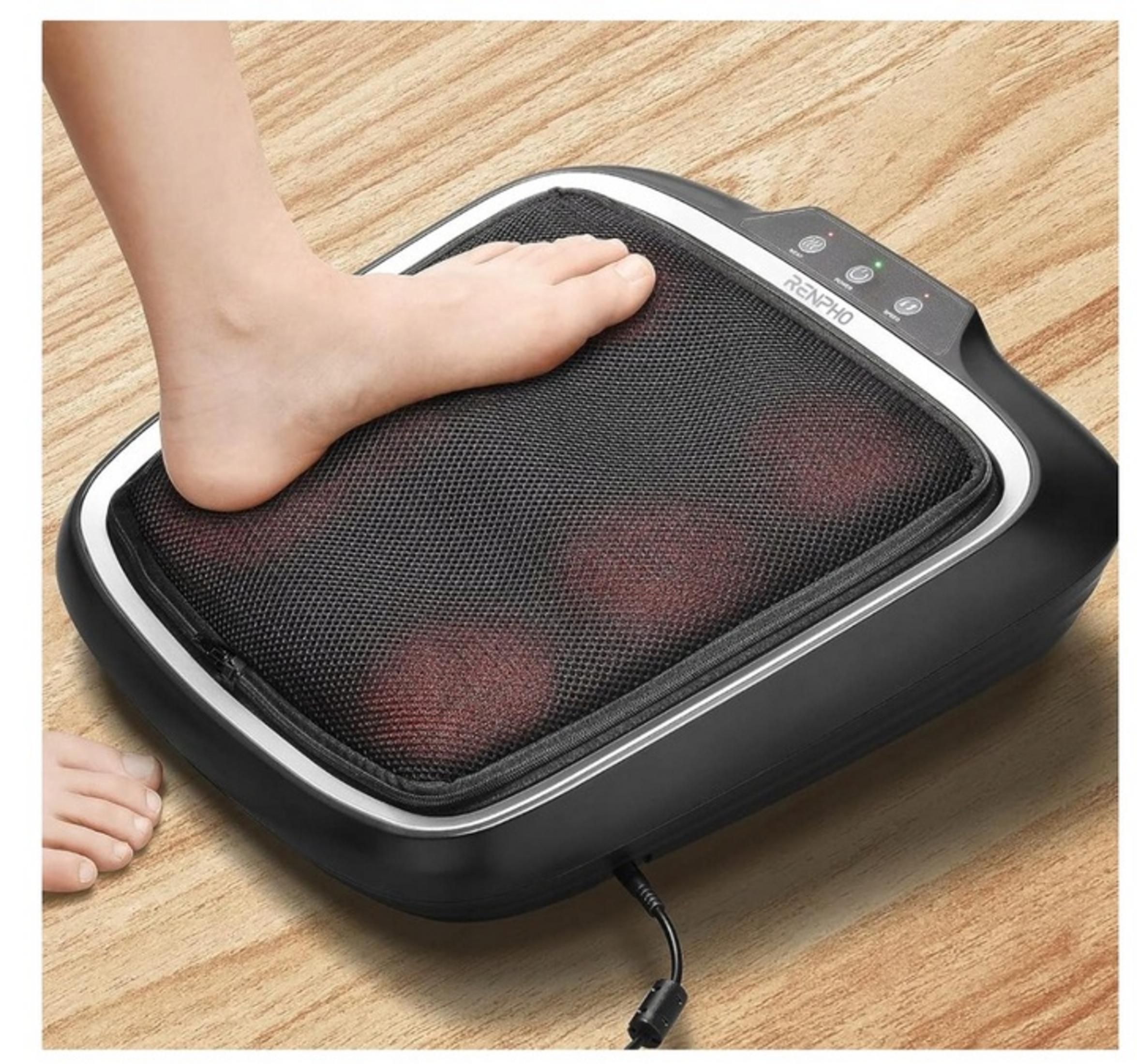 Ein Fuß ruht auf einem schwarzen Fußmassagegerät mit roten Lichtern auf Holzboden.