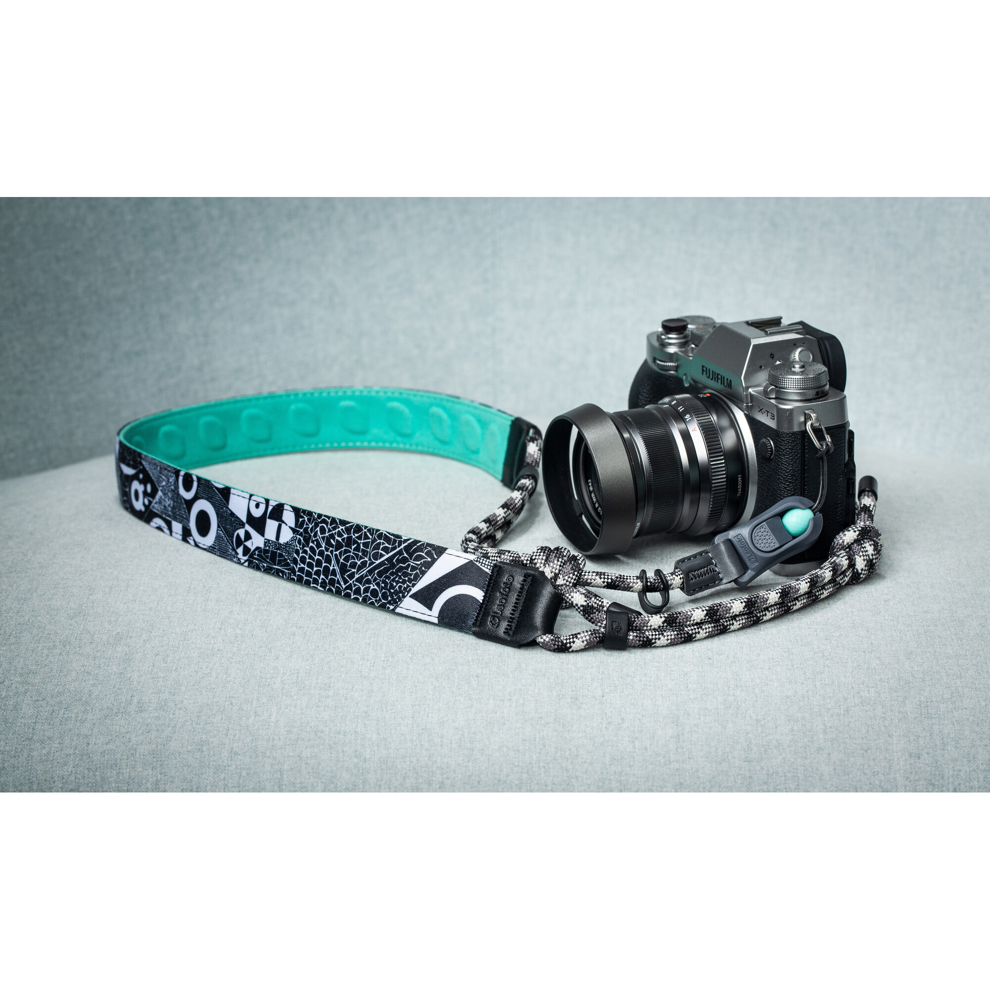 Una cámara Fujifilm plateada con correa negra y forro turquesa descansa sobre una superficie azul claro.