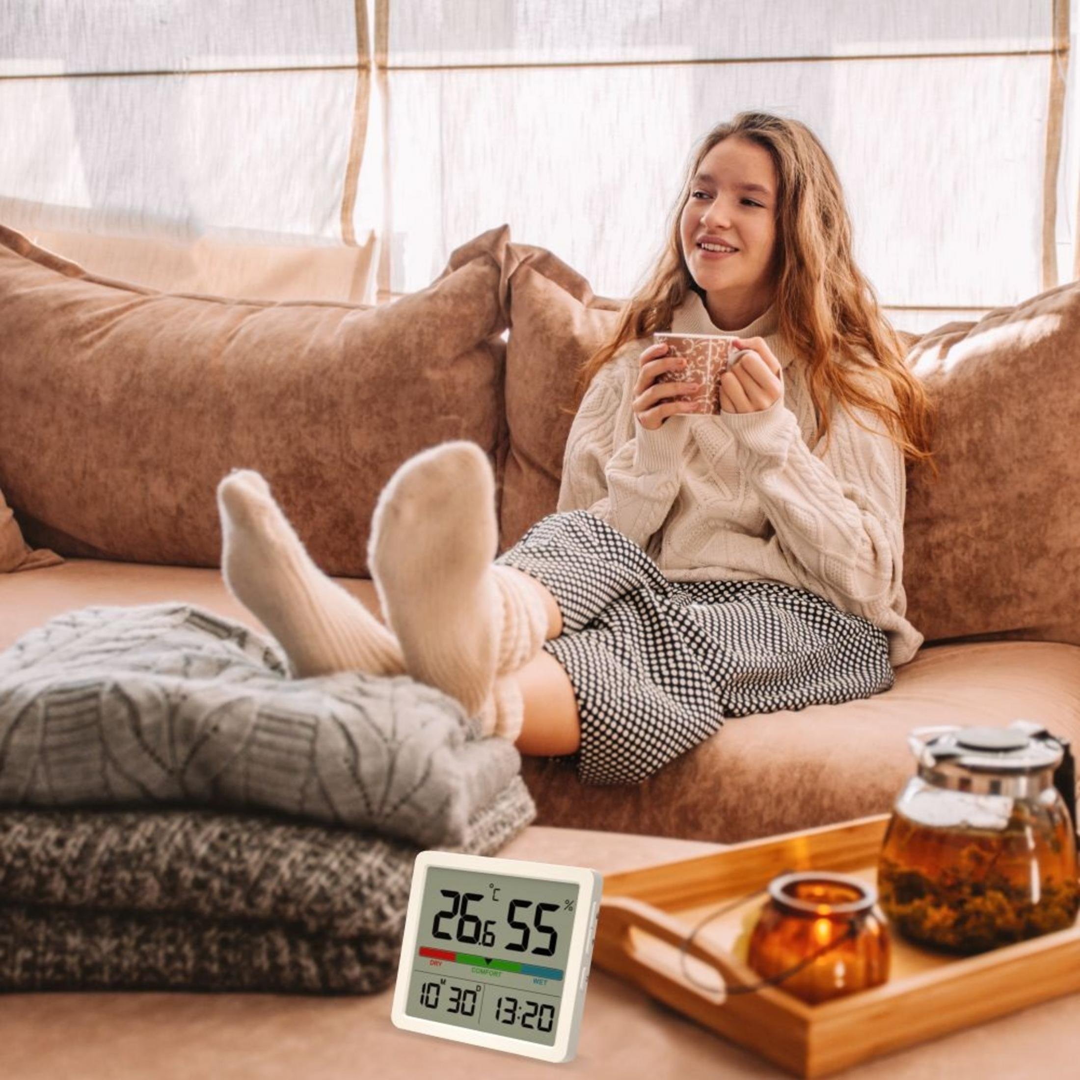Kobieta relaksuje się na kanapie z kubkiem, podczas gdy cyfrowy termometr pokazuje 26,6°C i 55% wilgotności.