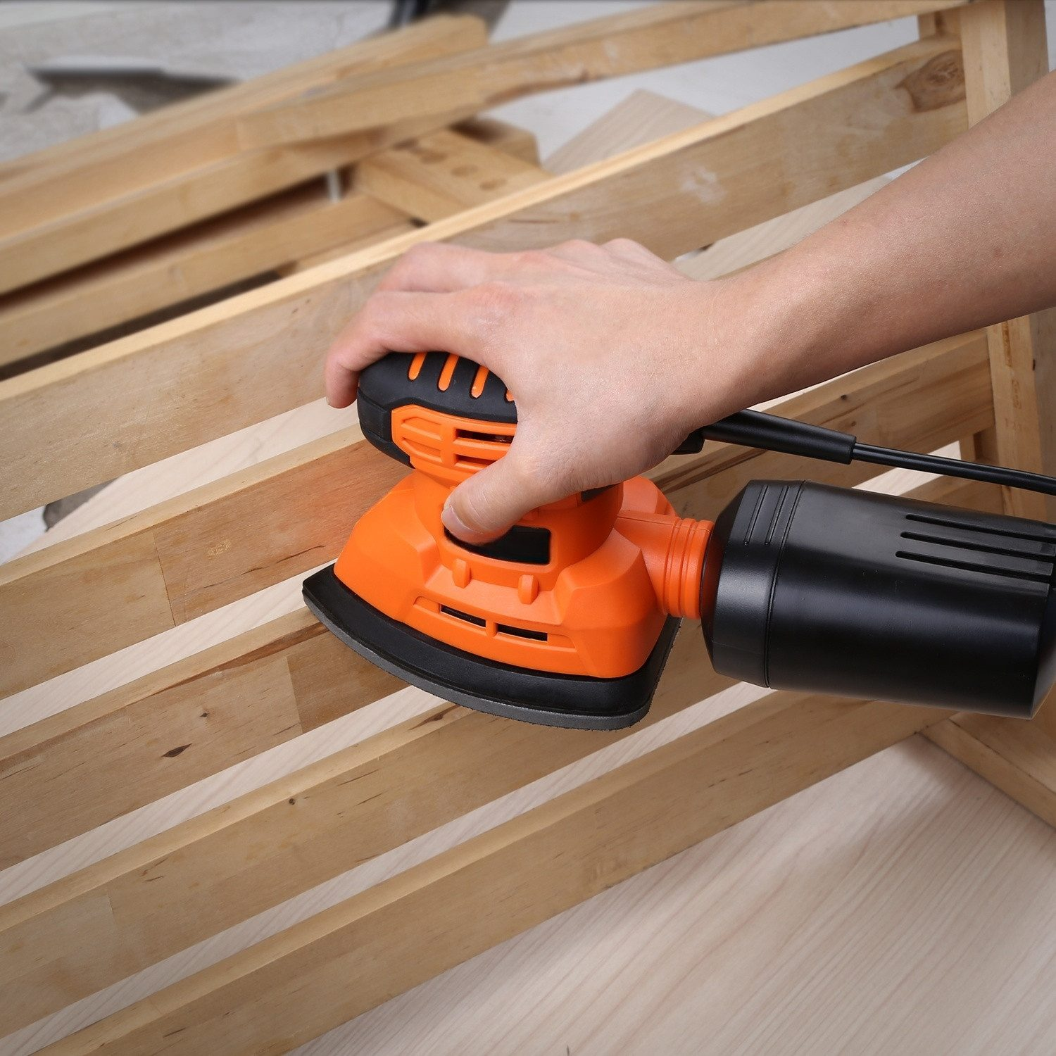 Hand benutzt einen orangefarbenen Exzenterschleifer auf hellen Holzlatten. Staubabscheider angeschlossen.