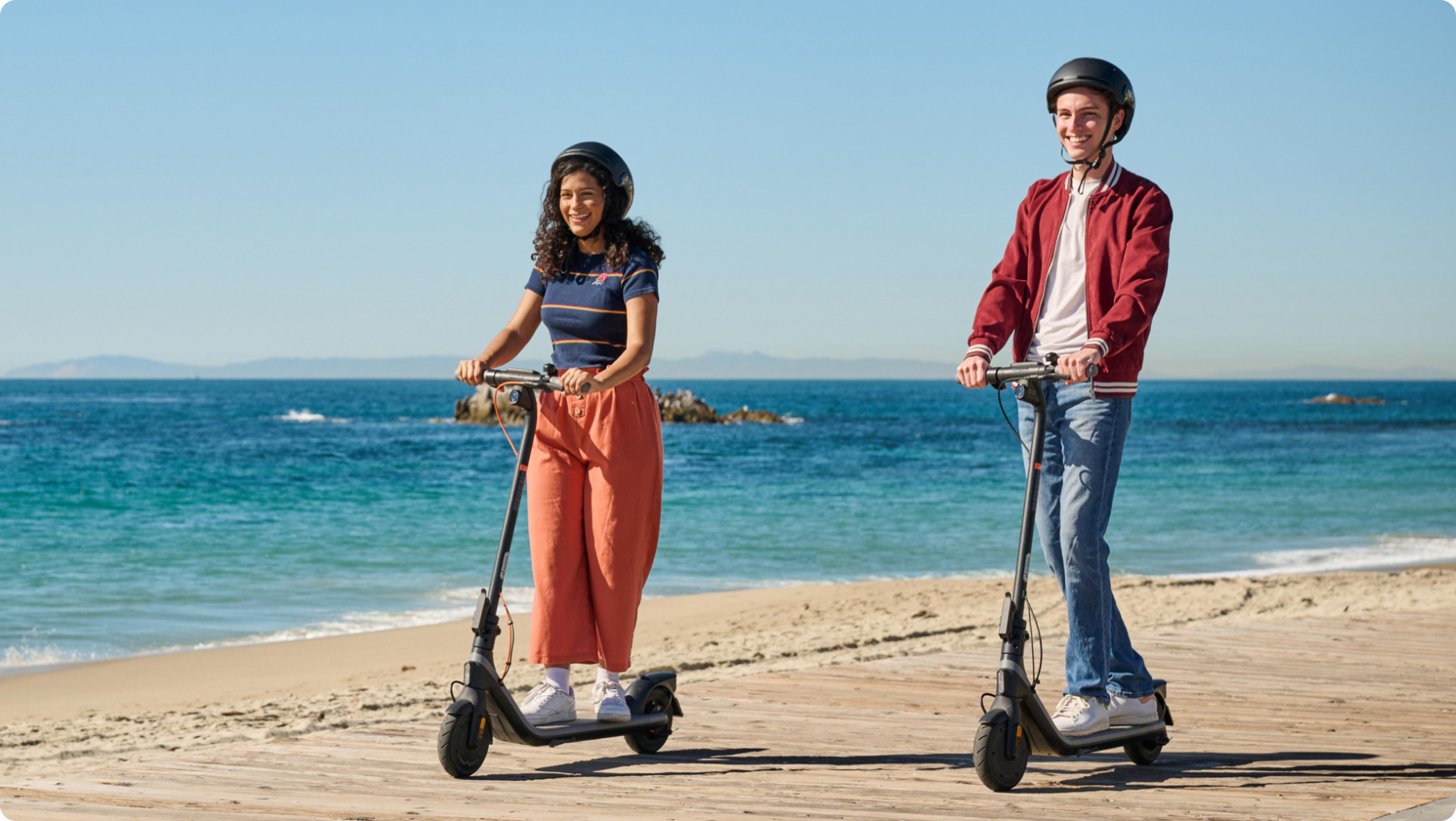 Dos personas montan patinetes eléctricos en un paseo marítimo junto al mar. Cielo despejado.