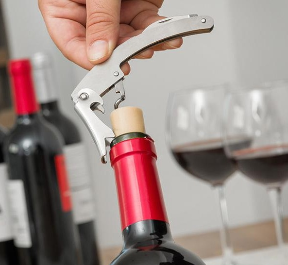 Una mano usa un sacacorchos para abrir una botella de vino tinto, con dos copas de vino cerca.