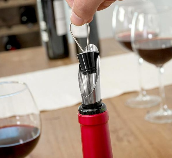 Una mano inserta un aireador de vino en una botella de vino tinto, con copas cerca.