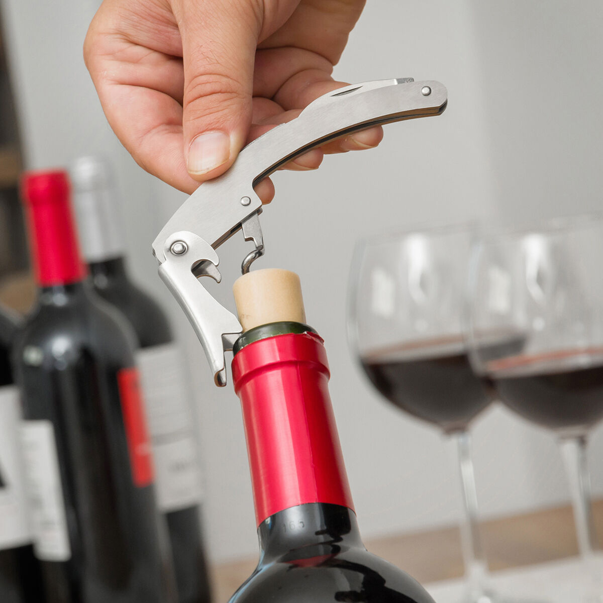 Una mano usa un sacacorchos para abrir una botella de vino tinto, con dos copas de vino cerca.