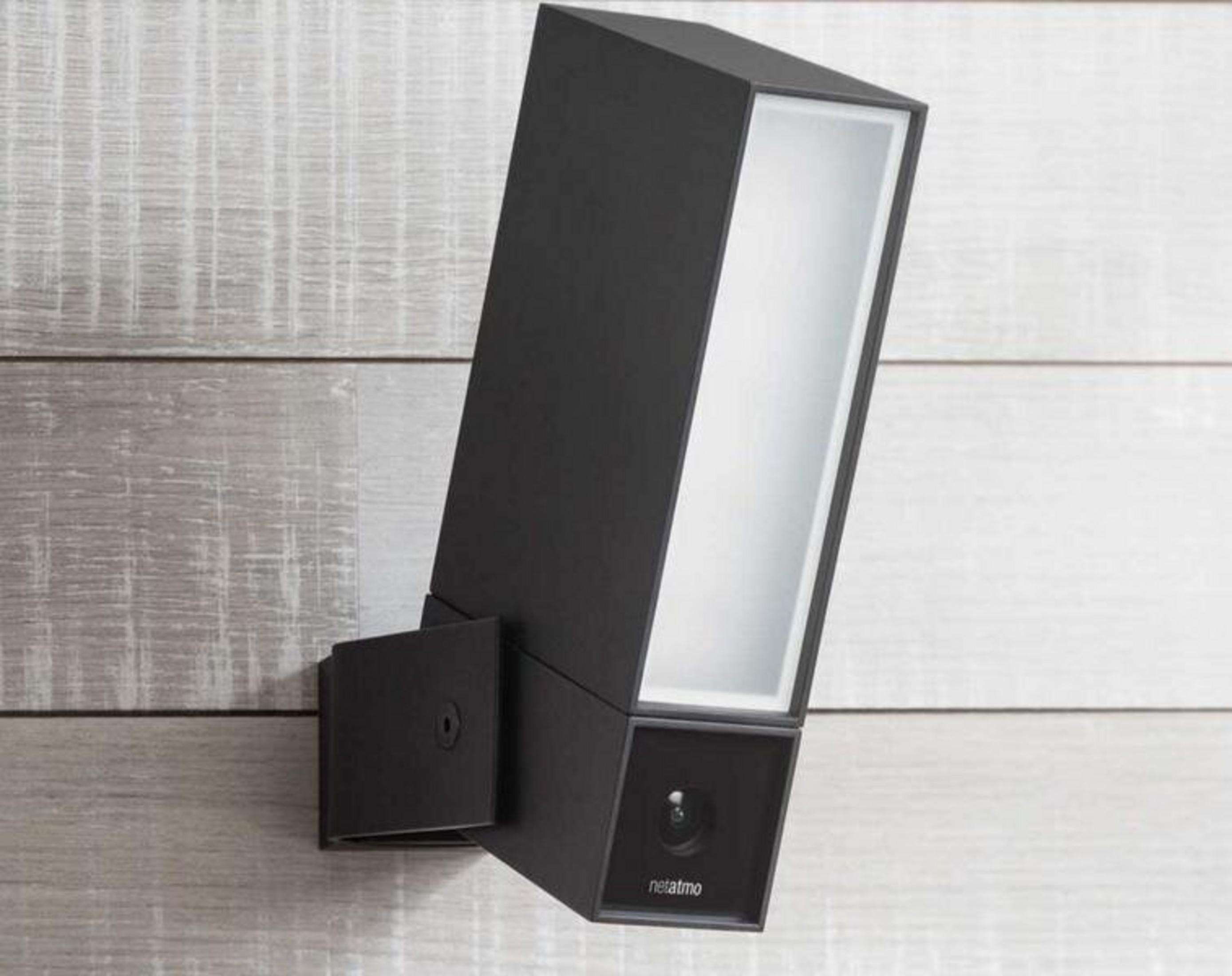 Una cámara de seguridad exterior Netatmo negra con luz está montada en una pared de madera gris claro.