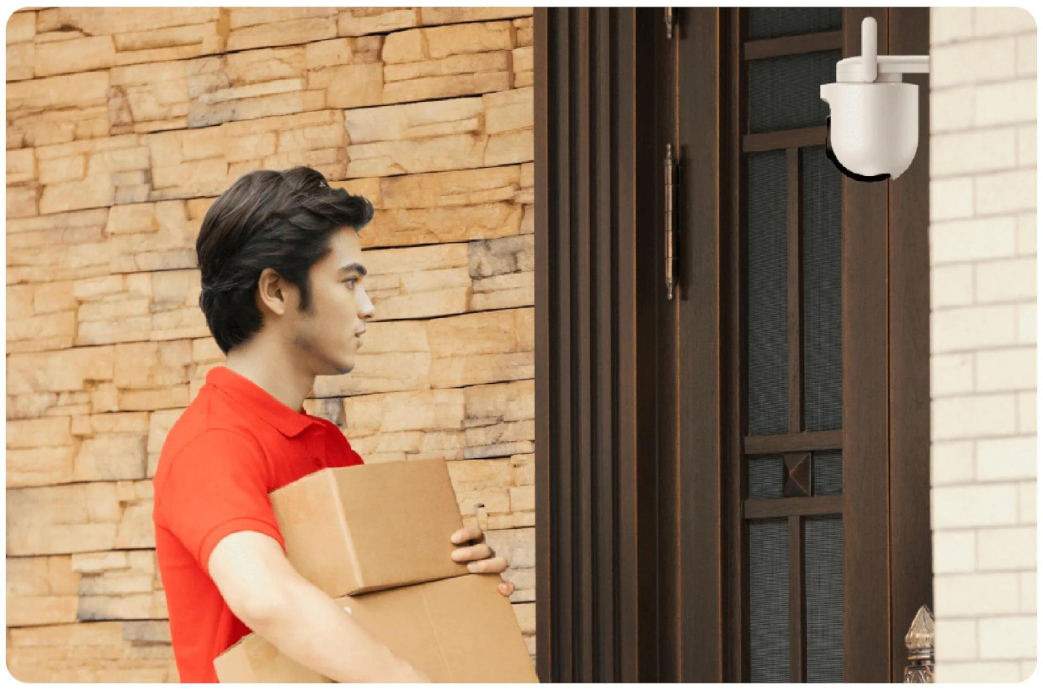 Listonosz w czerwonej koszulce niesie dwa brązowe kartony do drzwi z kamerą bezpieczeństwa.