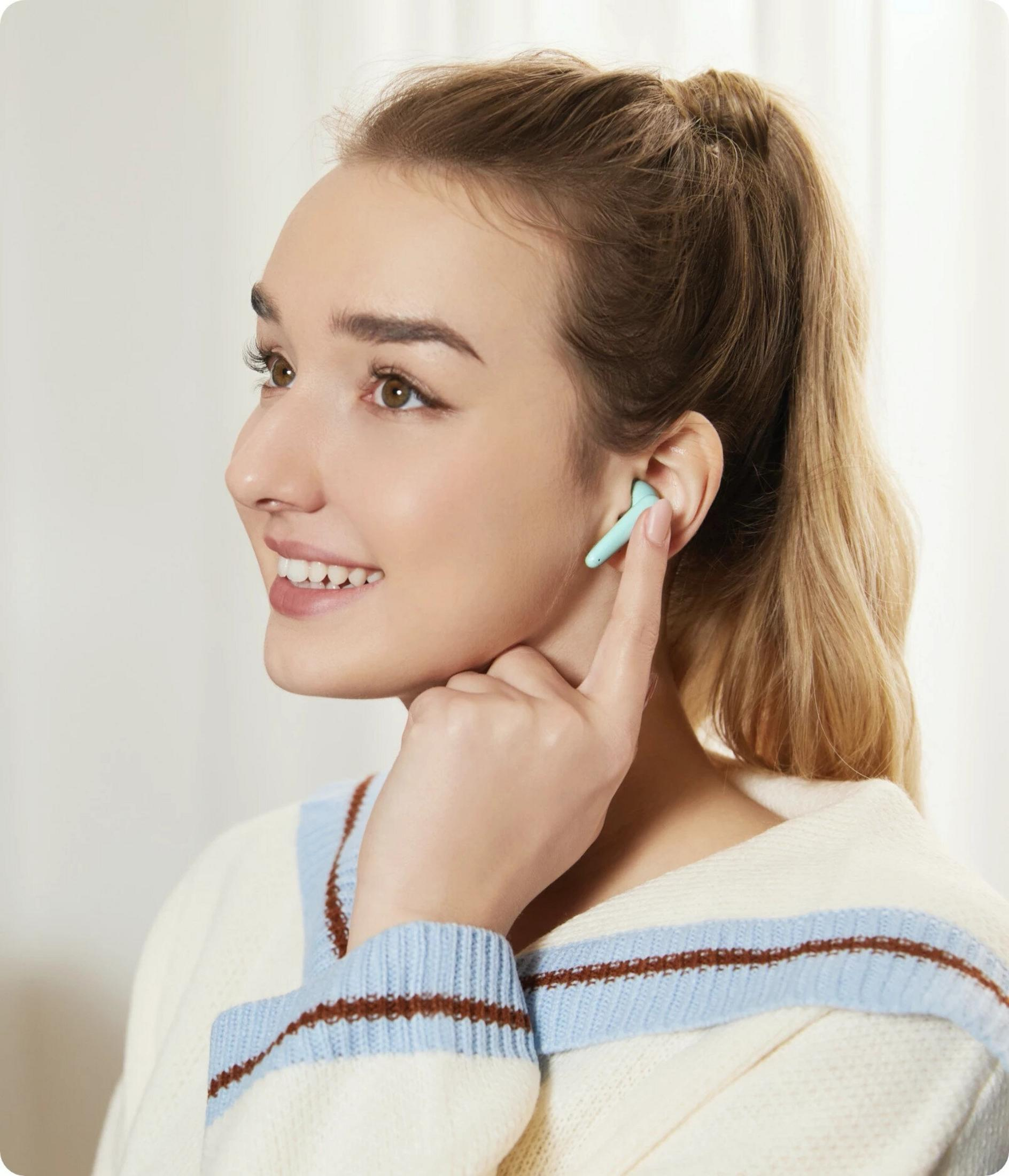 Una giovane donna con coda di cavallo bionda indossa un maglione bianco a righe blu e marroni e un auricolare verde menta.