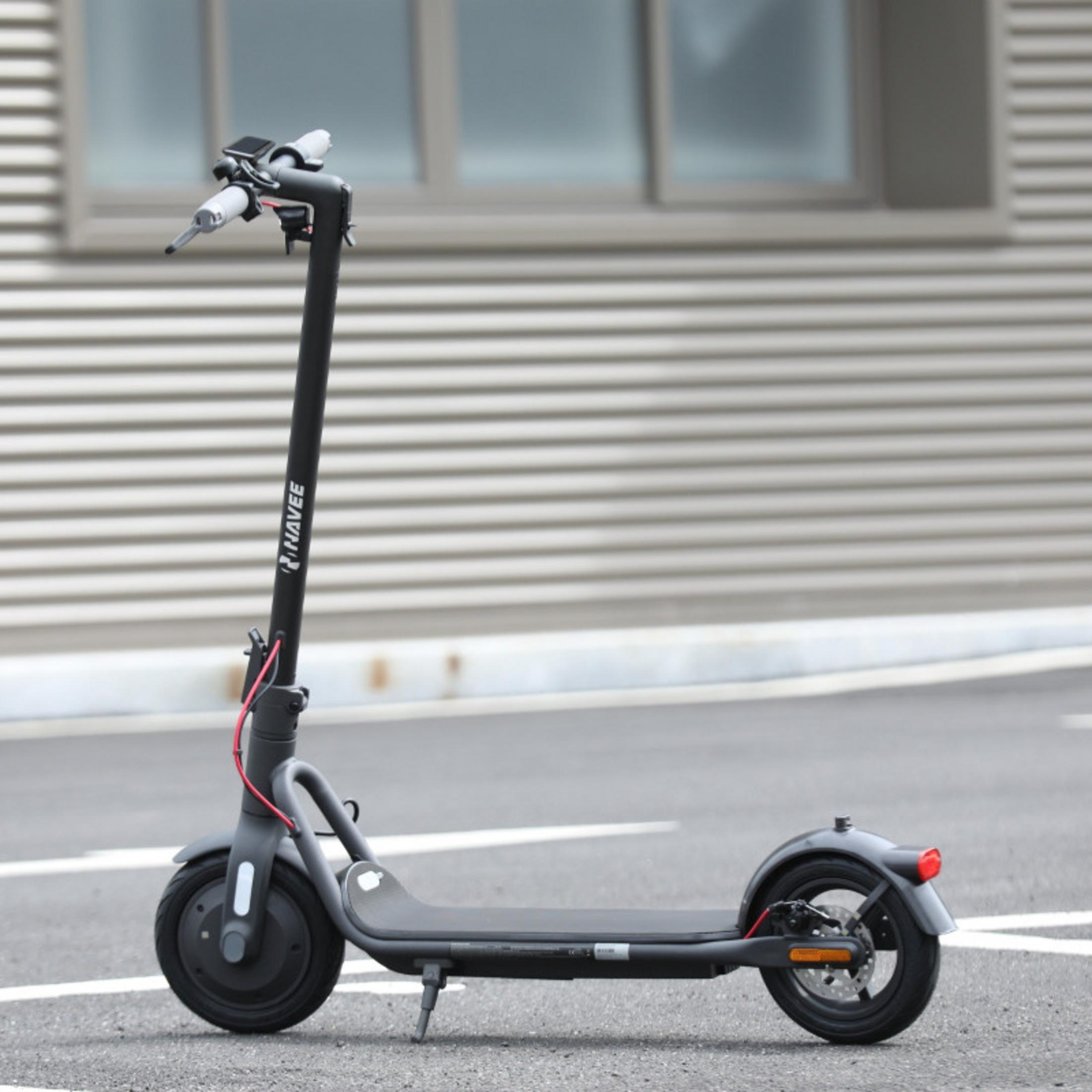 Ein dunkelgrauer Elektroroller steht auf Asphalt mit weißen Linien. Gebäude im Hintergrund.