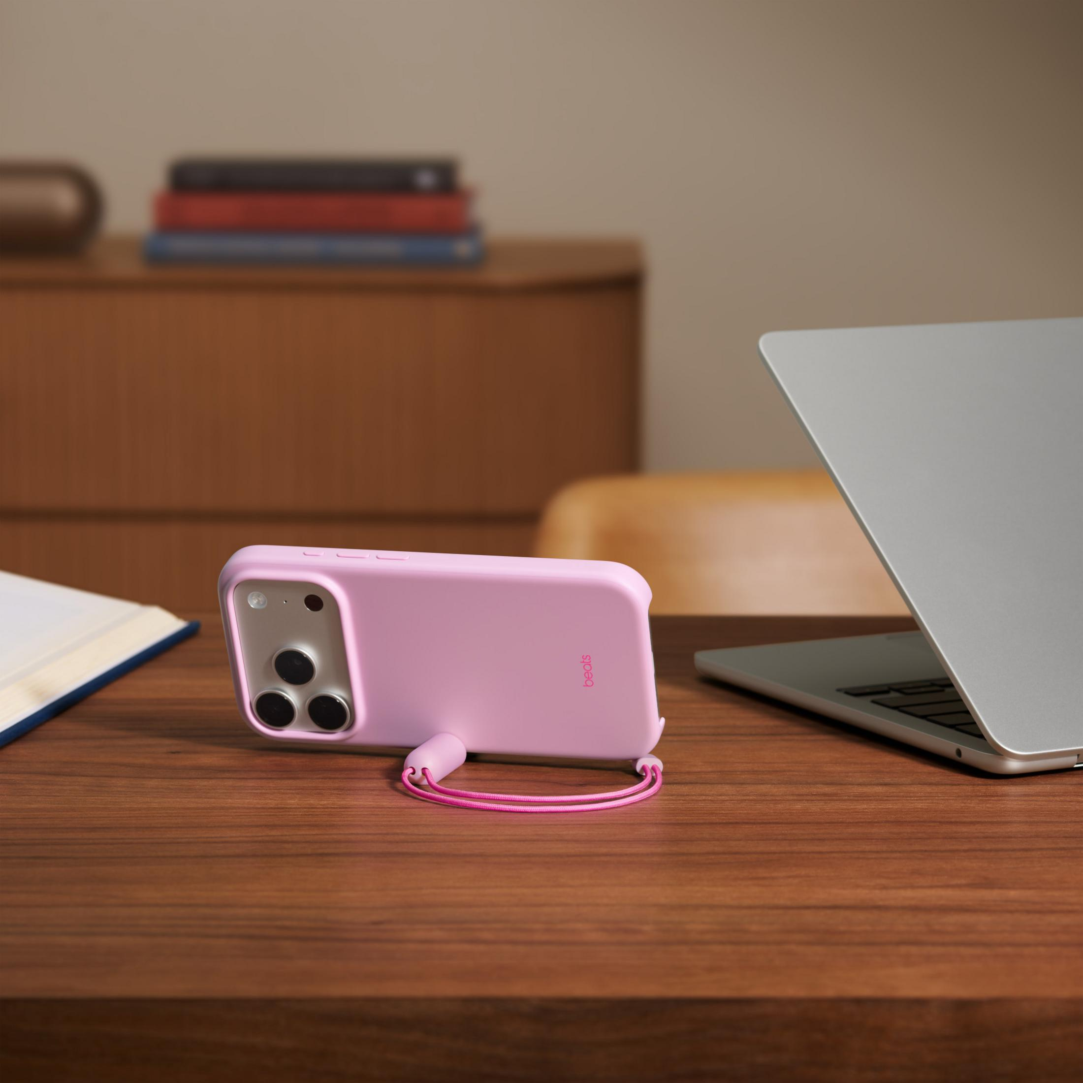 Una funda de teléfono rosa con un lazo se sienta en un escritorio de madera junto a un portátil y un libro.