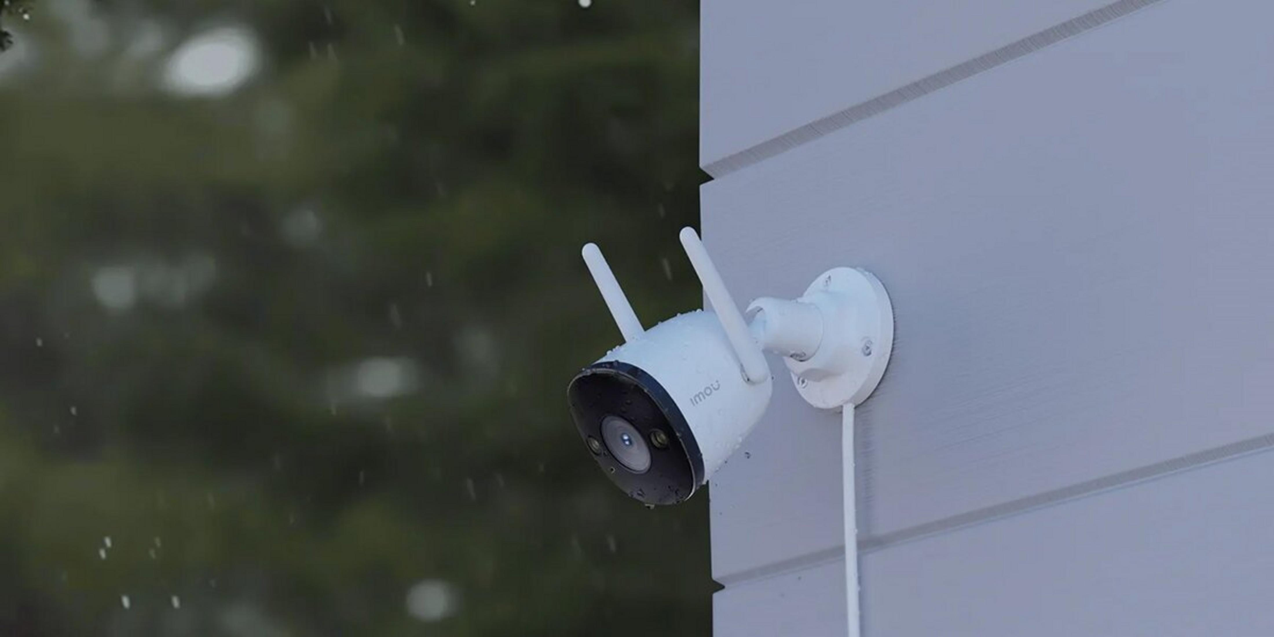 Een witte beveiligingscamera met antennes is gemonteerd op een lichtgrijze muur, er valt regen.