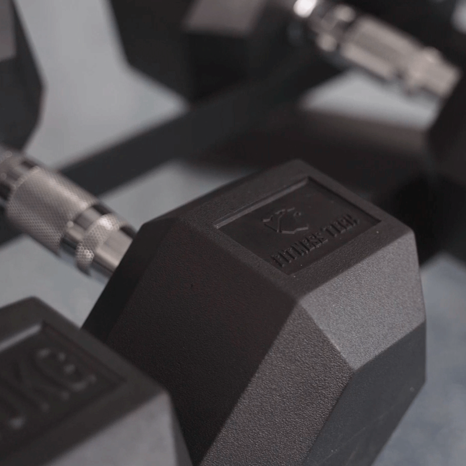 Mancuernas hexagonales negras en un rack. Una mancuerna tiene el logo 'Fitness Tech'.