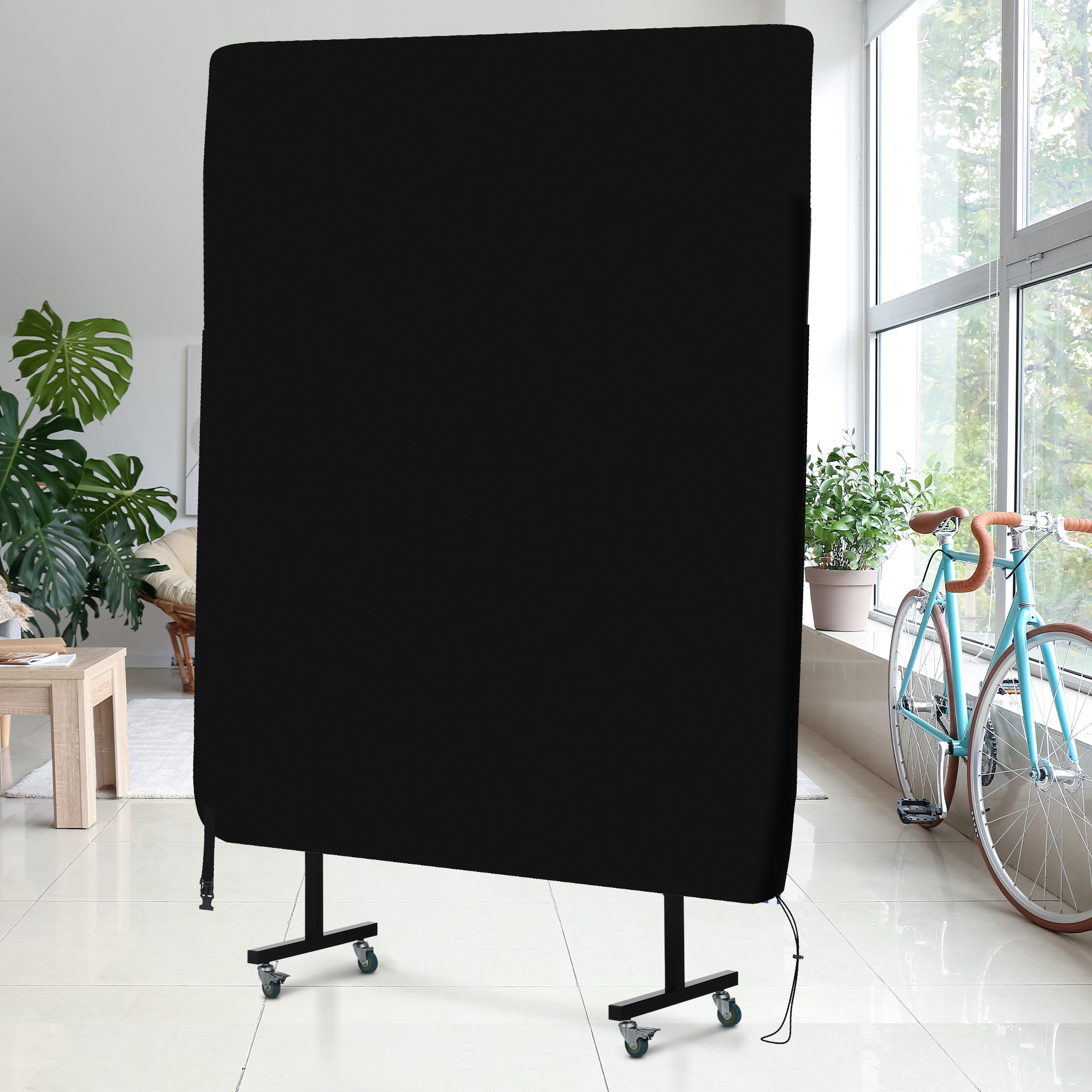 Una pantalla negra grande con ruedas se encuentra en una habitación con plantas y una bicicleta azul junto a una ventana.