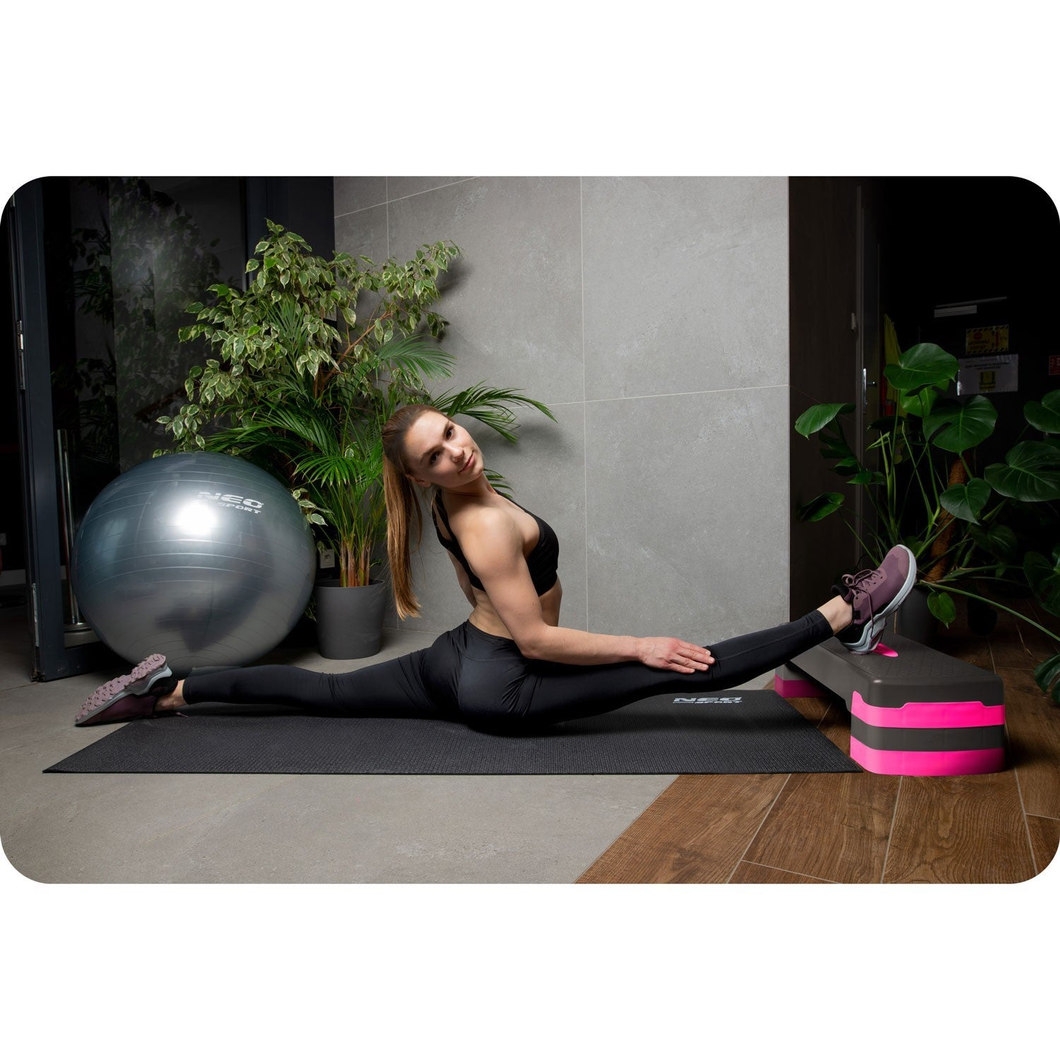 Mujer en ropa deportiva negra haciendo el spagat en esterilla, con balón de ejercicio y plantas.