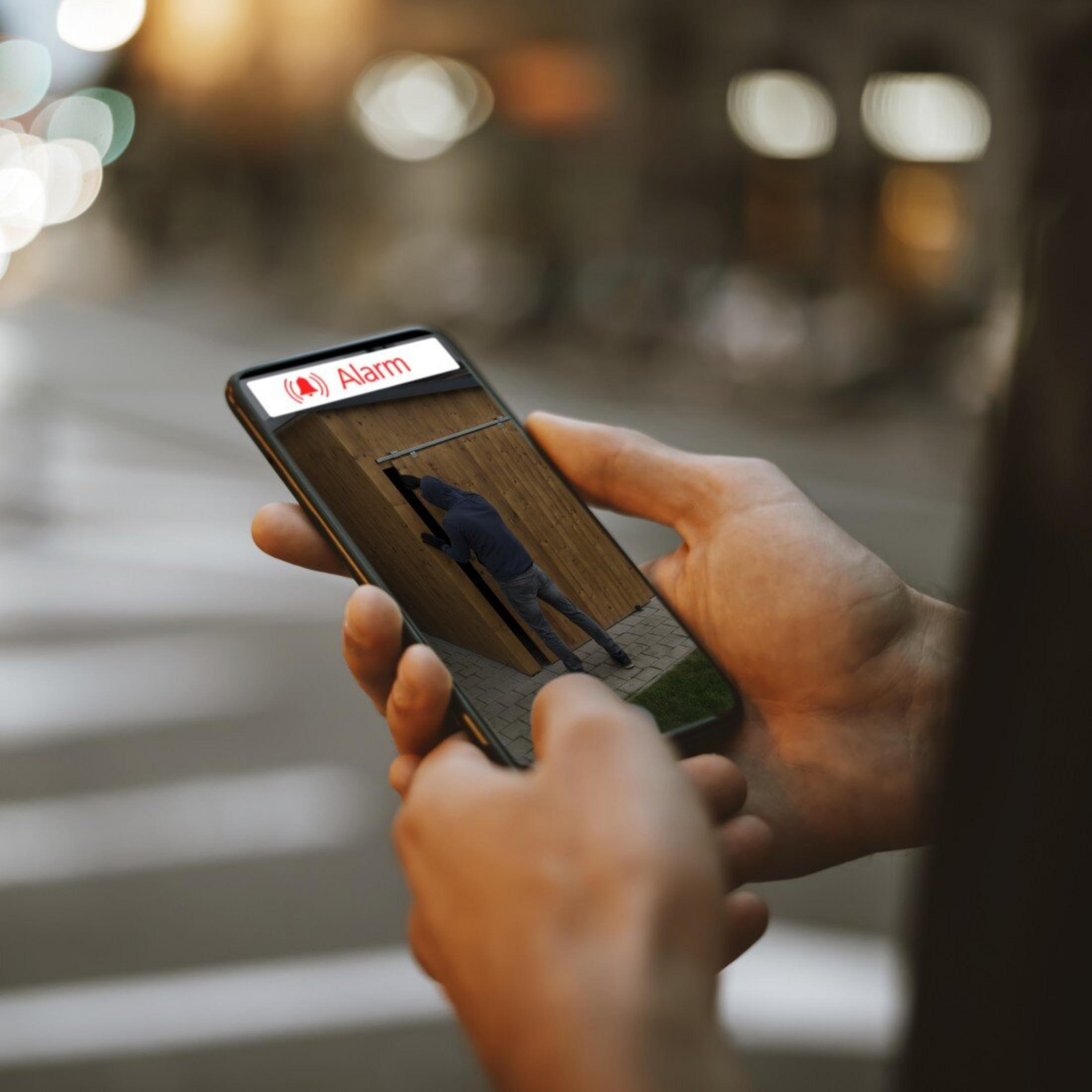 Osoba w ciemnym ubraniu wspina się po drewnianym płocie. Wyświetlany jest alarm.