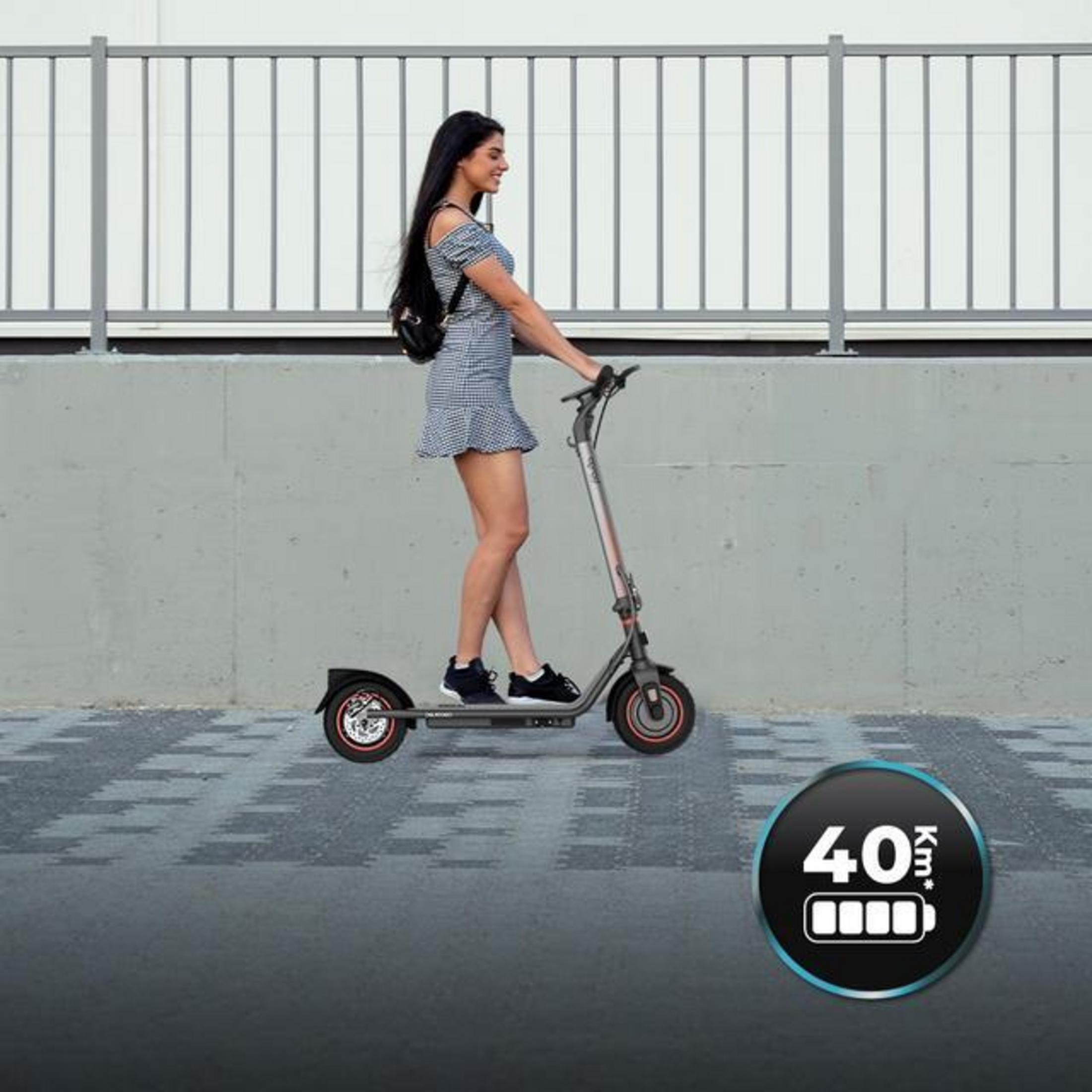 Frau auf Elektroroller, graue Betonwand, karierter Boden, 40km Akku-Symbol.