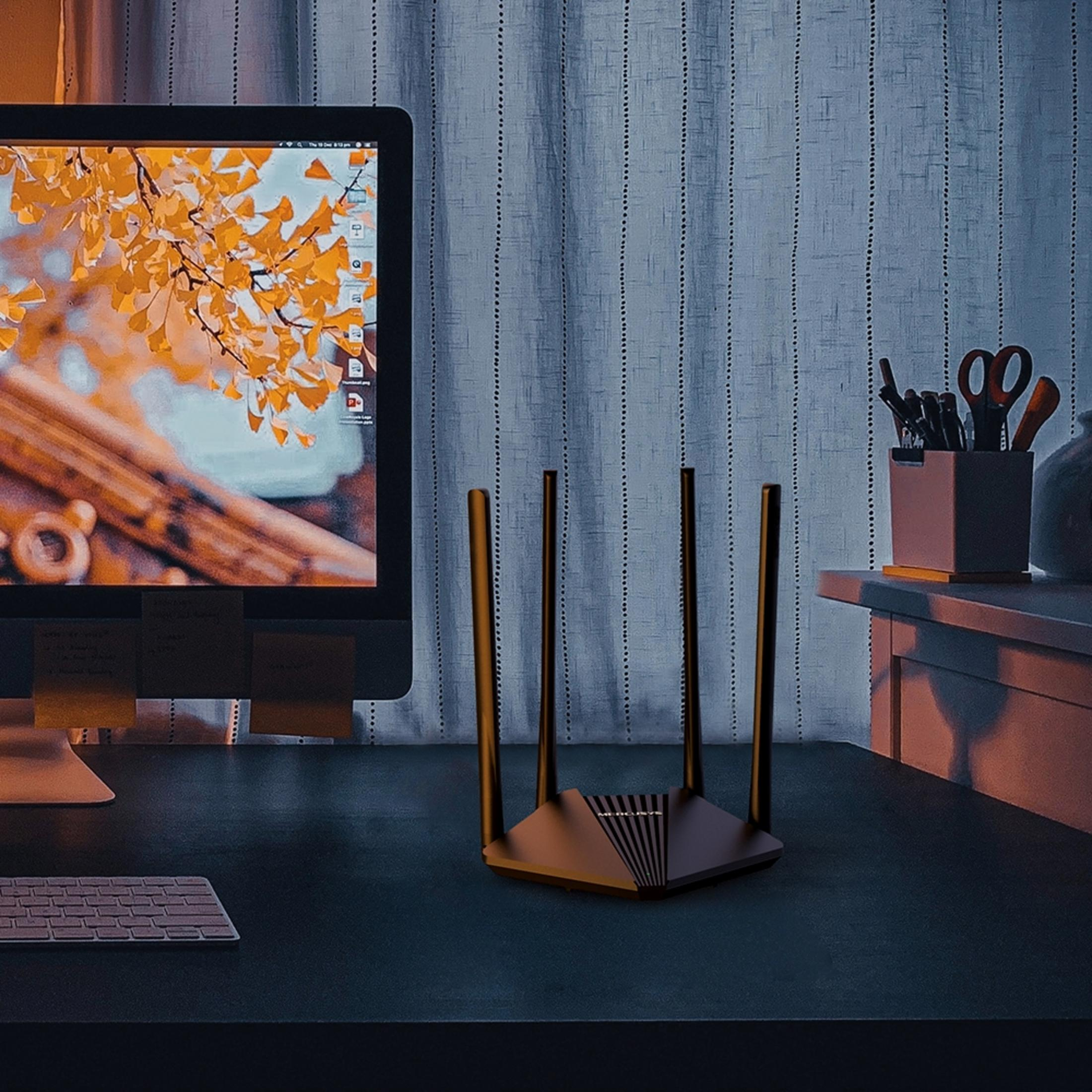 Czarny router z czterema antenami stoi na biurku obok monitora komputerowego wyświetlającego jesienne liście.