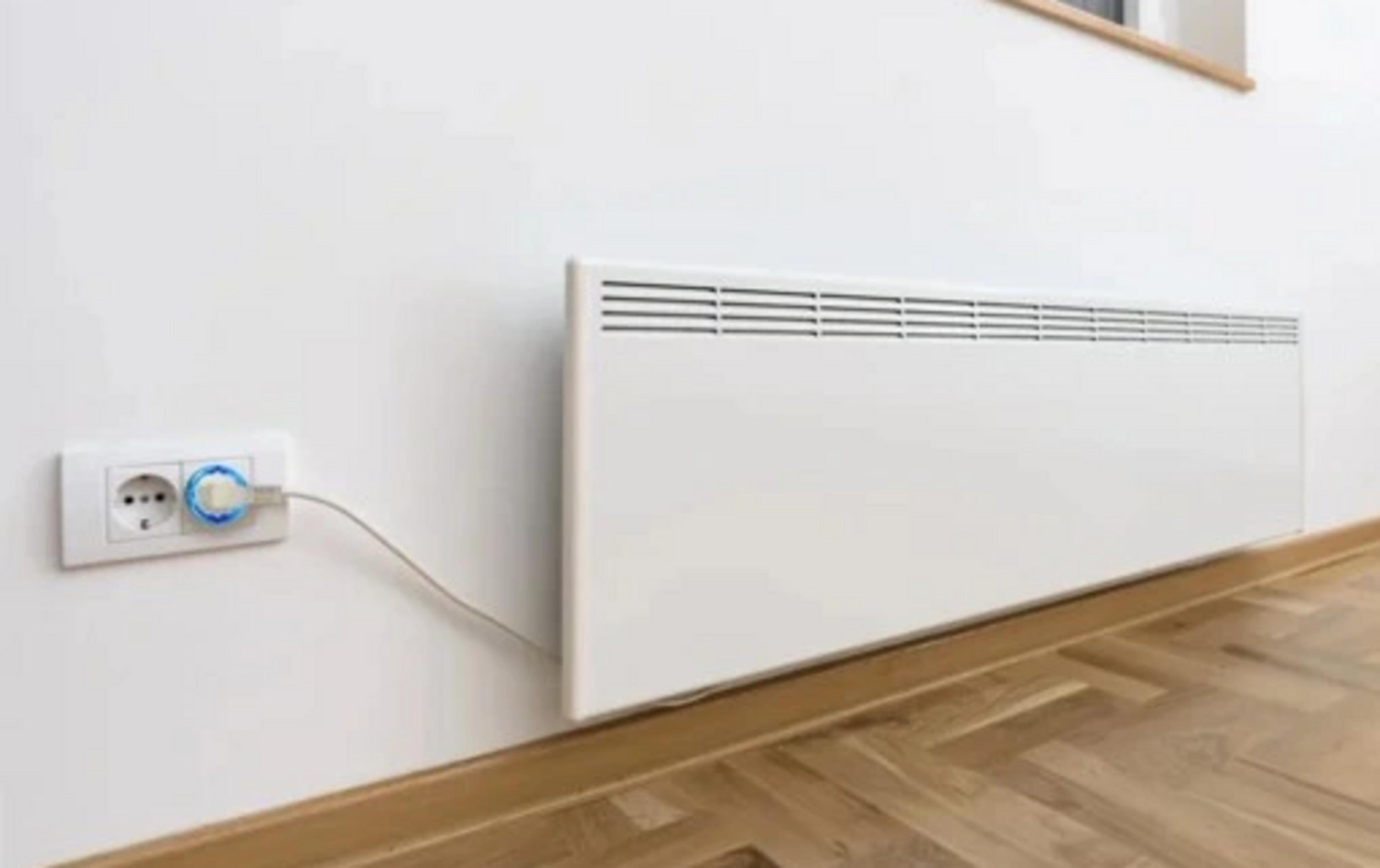 Biały grzejnik konwektorowy na białej ścianie, obok gniazdko elektryczne z wtyczką.