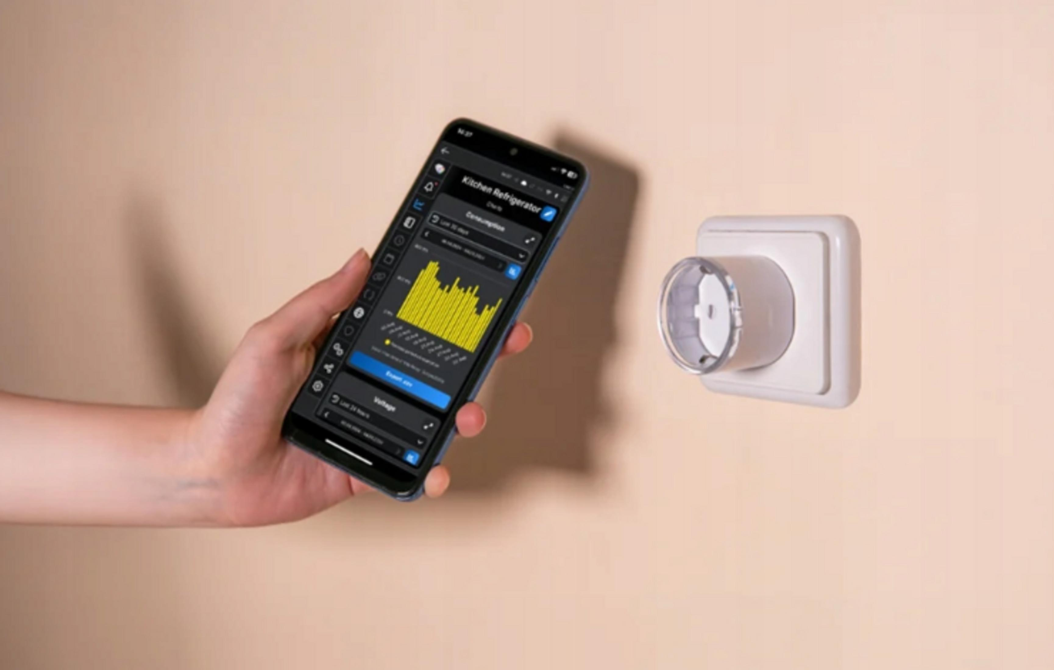 Dłoń trzyma smartfona pokazującego dane o zużyciu energii obok gniazdka ściennego.
