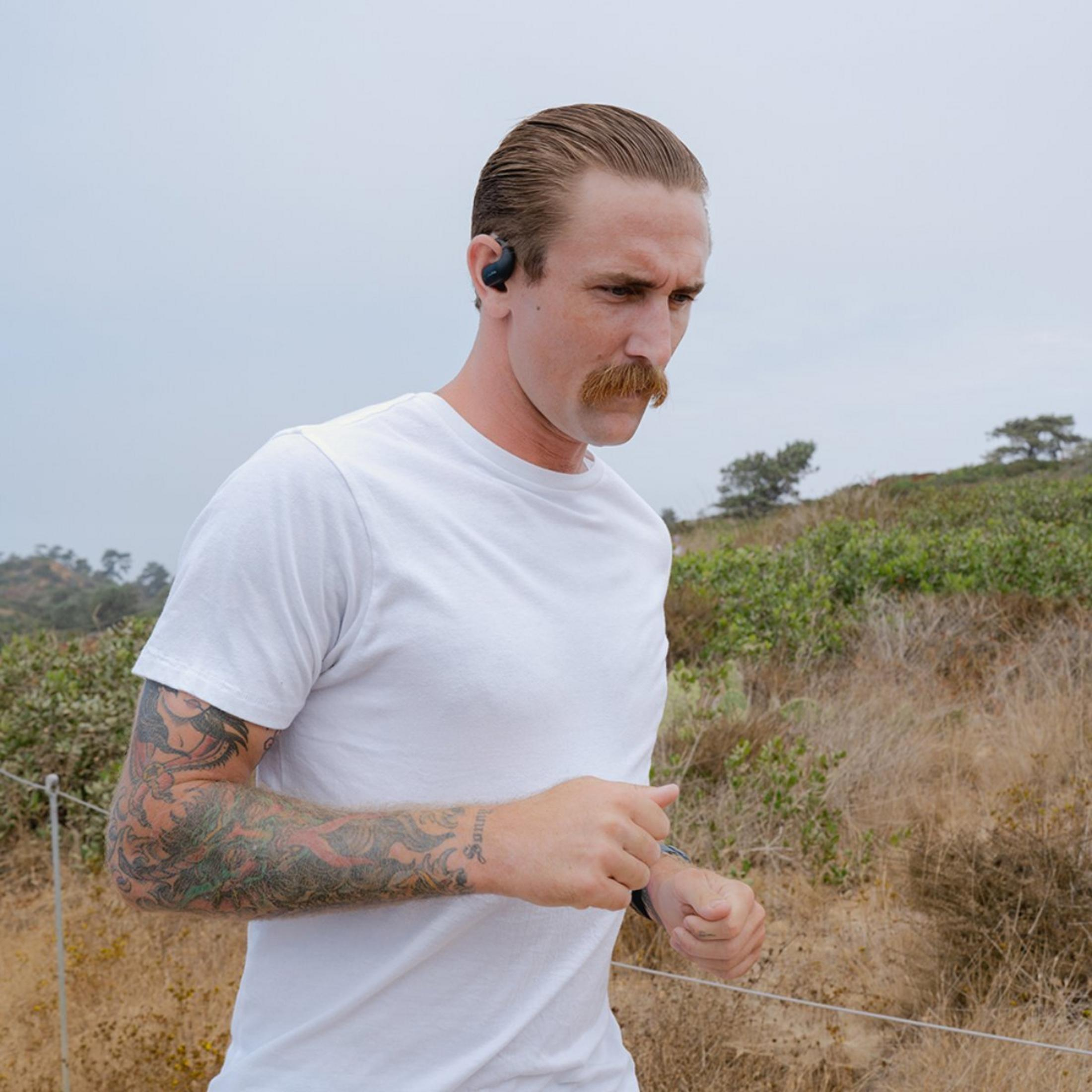 Homme avec moustache et tatouages court dehors avec écouteurs. Paysage sec et herbeux.