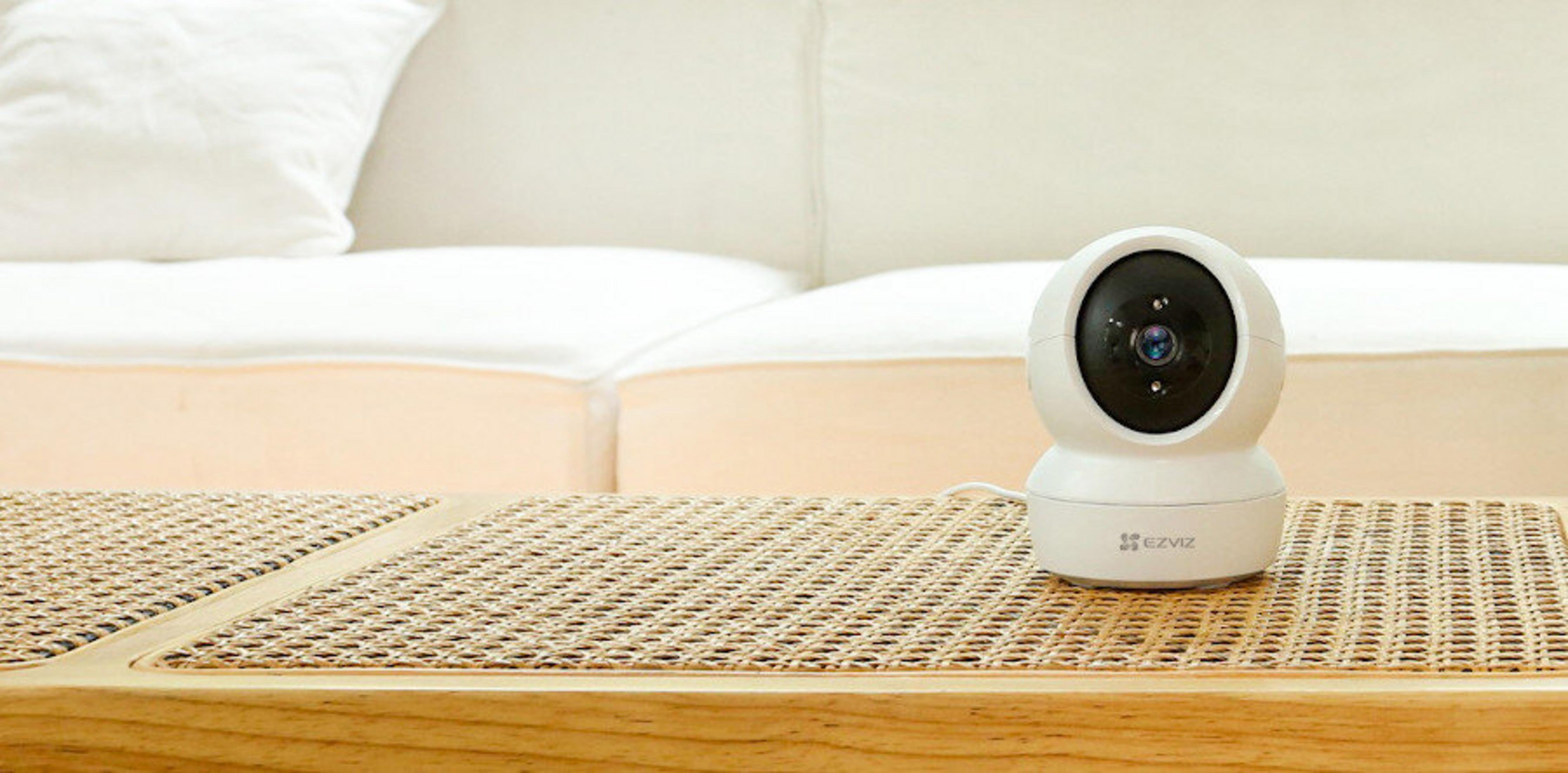 Biała kamera EZVIZ stoi na plecionej ławie przed kanapą.