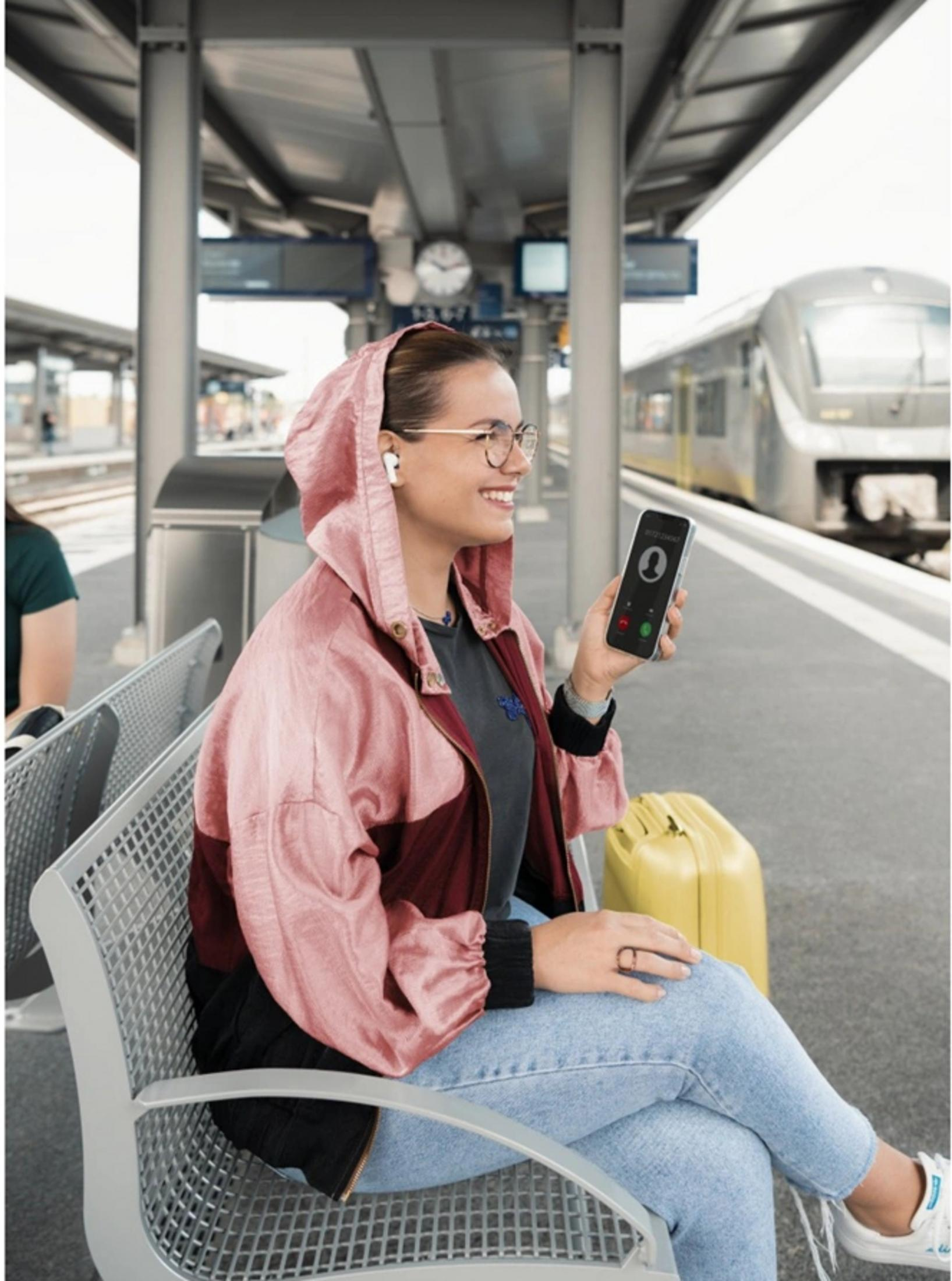 Una donna con auricolari siede sulla banchina di una stazione ferroviaria tenendo uno smartphone.