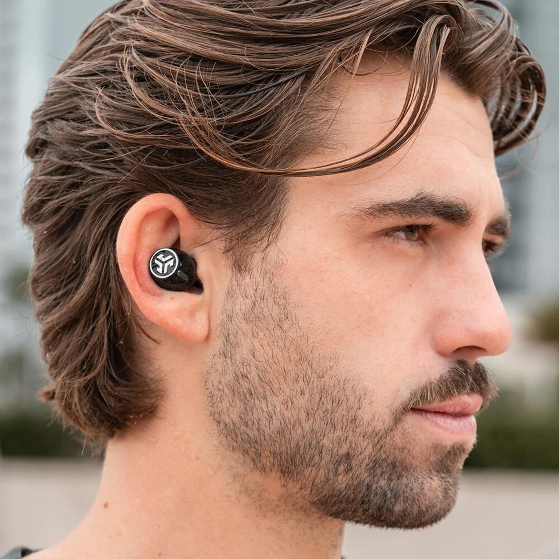 Primo piano di un uomo con capelli castani e barba, che indossa un auricolare nero.