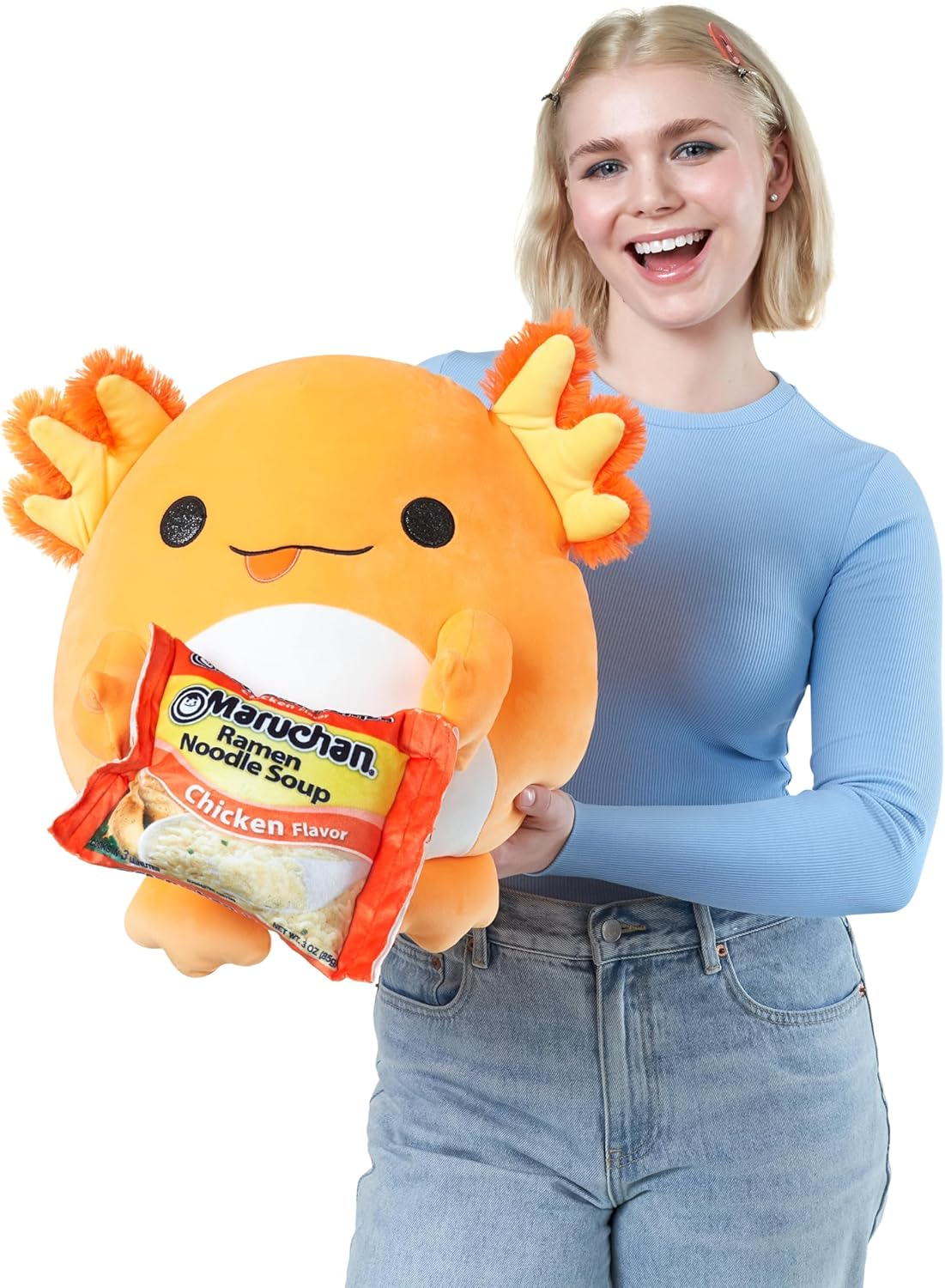 Eine lächelnde Frau hält ein orangefarbenes Axolotl-Plüschtier mit einer Packung Hühnerramen von Maruchan.