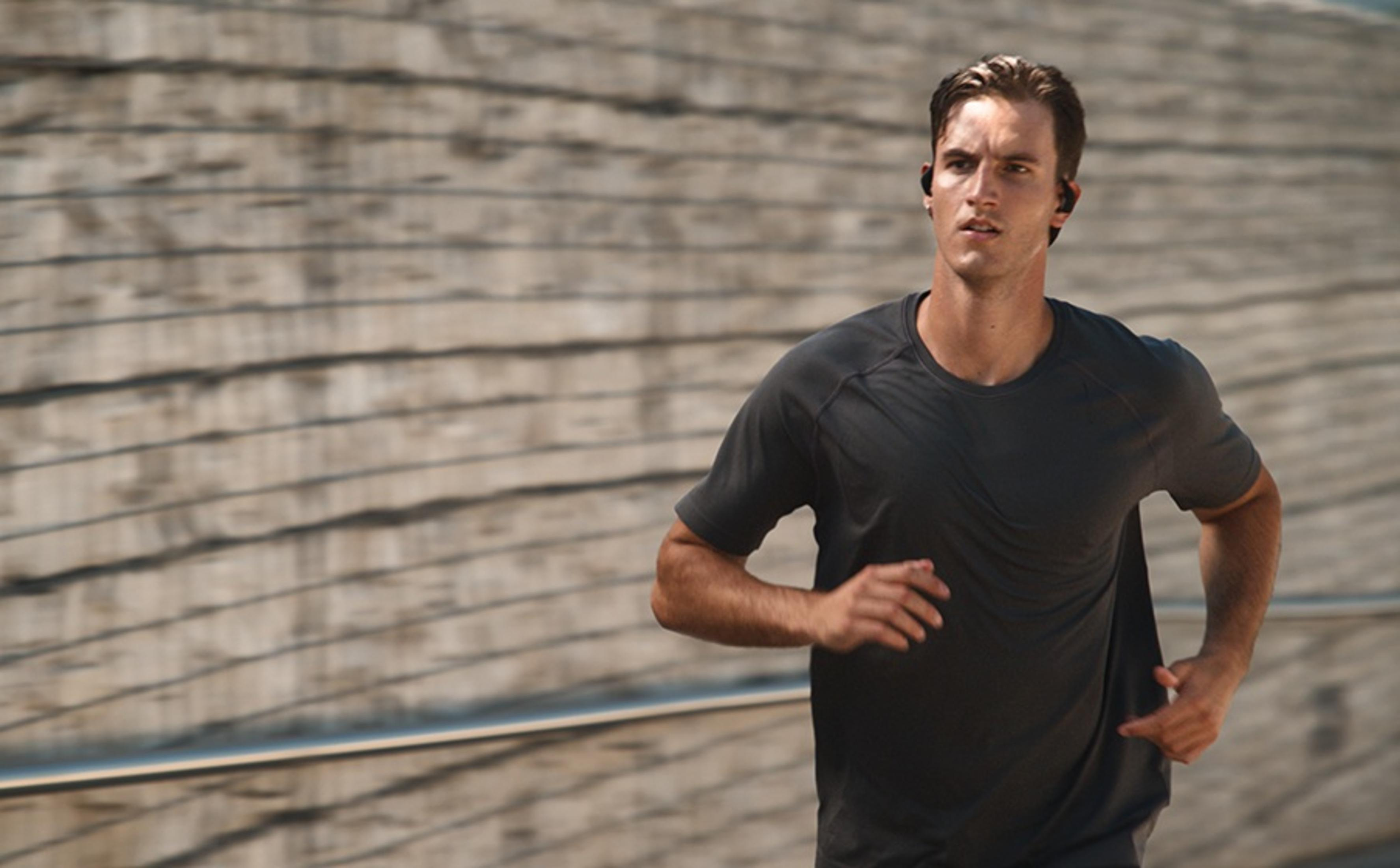 Mężczyzna biegnie na zewnątrz, w ciemnej koszulce i słuchawkach bezprzewodowych. Rozmyte tło.