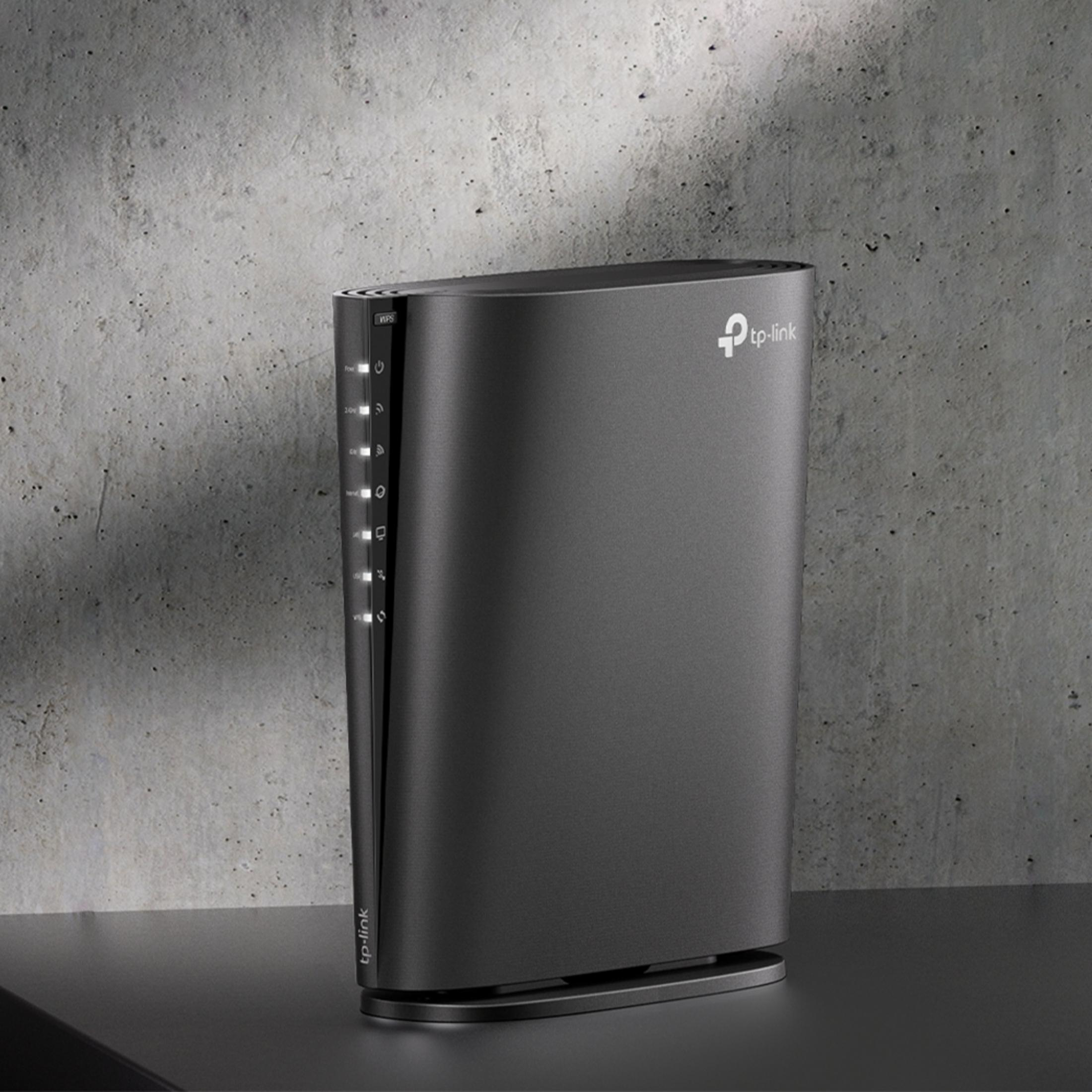 Czarny router TP-Link ze wskaźnikami świetlnymi stoi na ciemnej powierzchni na tle betonowej ściany.
