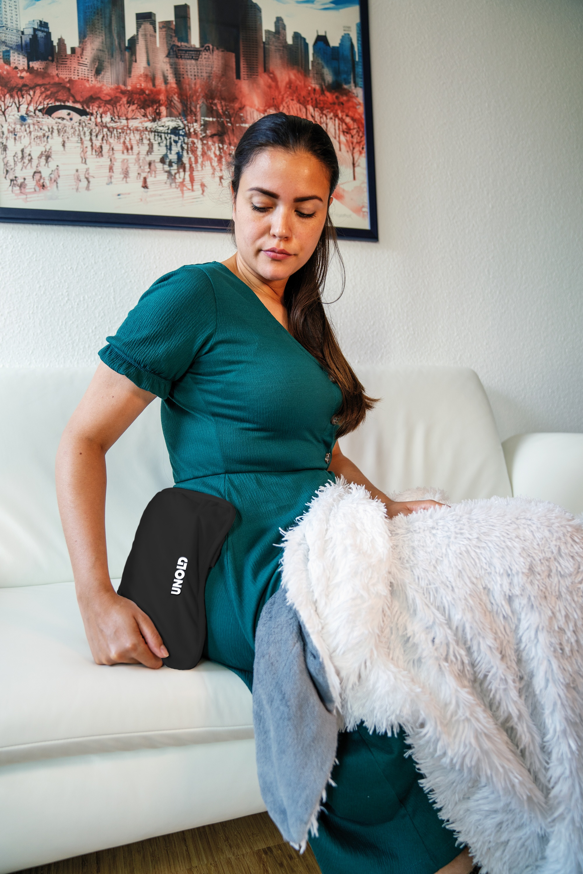 Eine Frau mit dunklem Haar sitzt auf einem weißen Sofa und hält ein schwarzes Heizkissen.