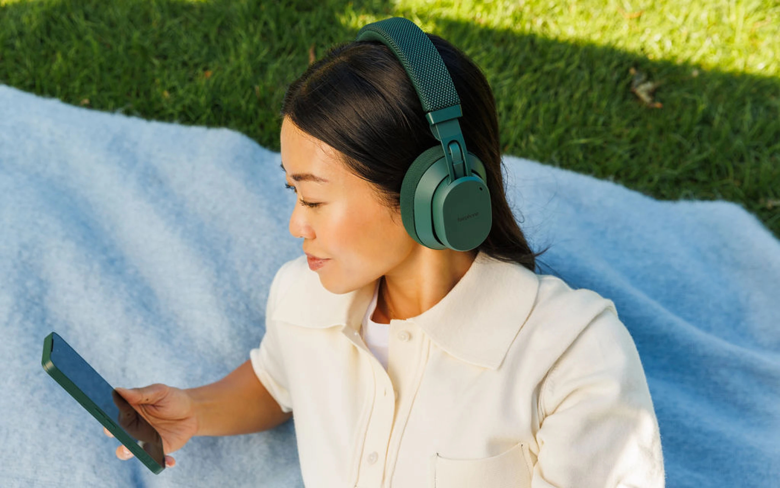 Eine Frau mit Kopfhörern liegt auf einer Decke im Gras und schaut auf ihr Handy.