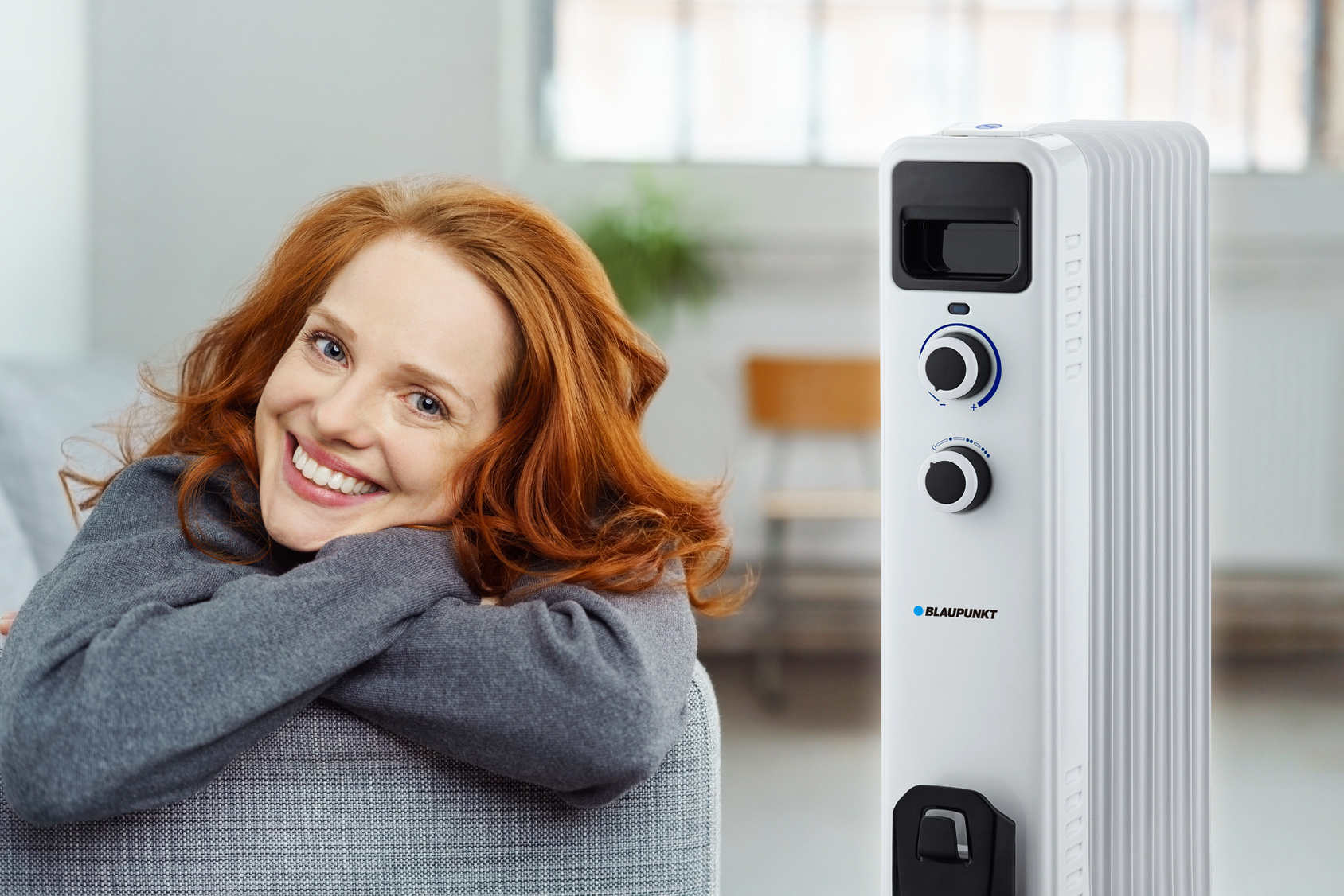 Kobieta z rudymi włosami uśmiecha się, opierając się o grzejnik olejowy.