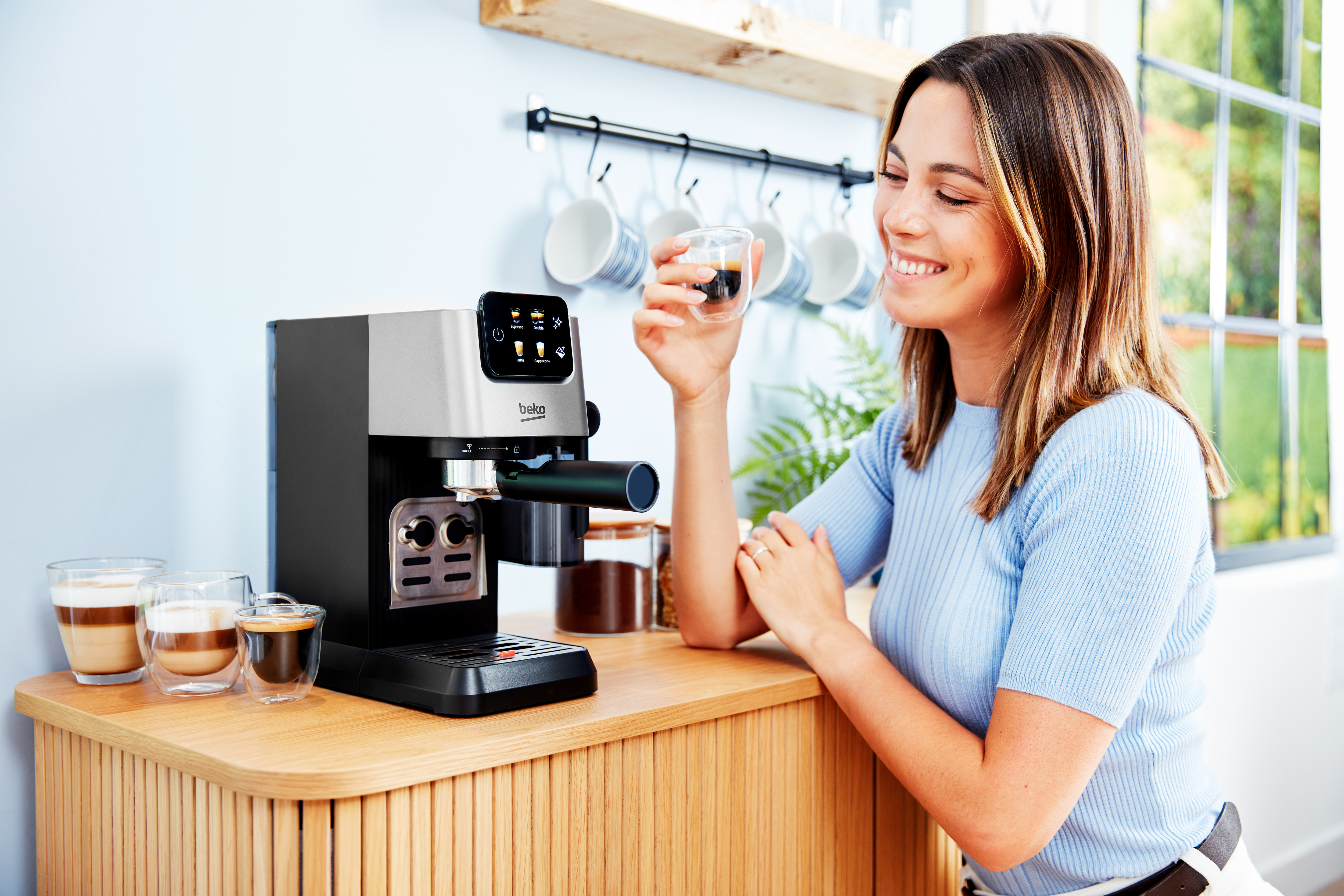 Eine Frau lächelt und hält eine kleine Espressotasse neben einer Beko-Kaffeemaschine.