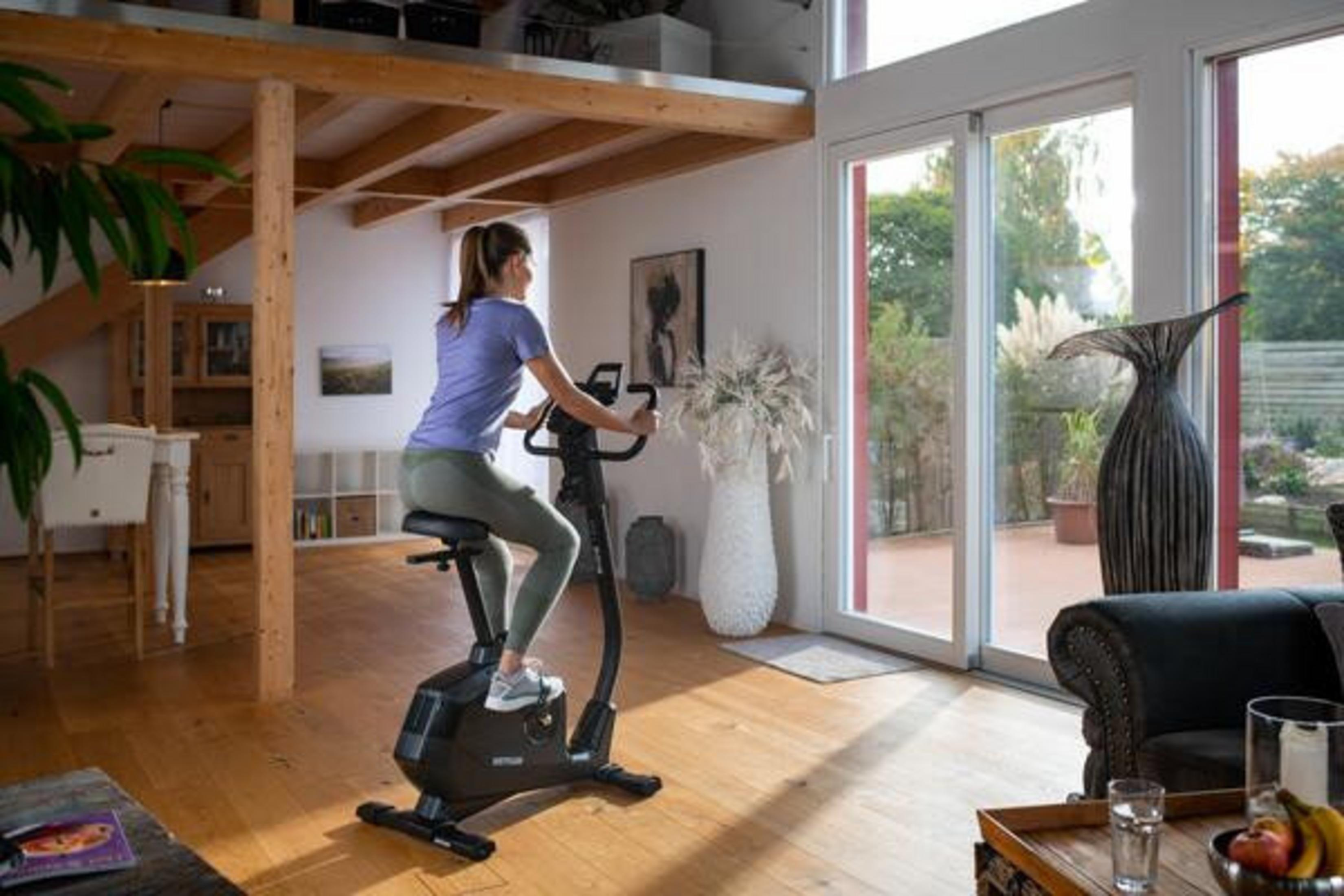 Mujer en bicicleta estática en interior, habitación luminosa con ventanas grandes.