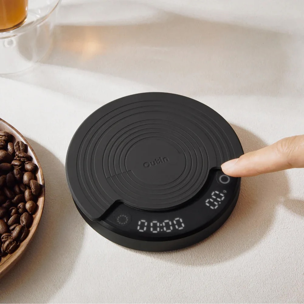 A finger presses the power button on a black digital coffee scale next to coffee beans.
