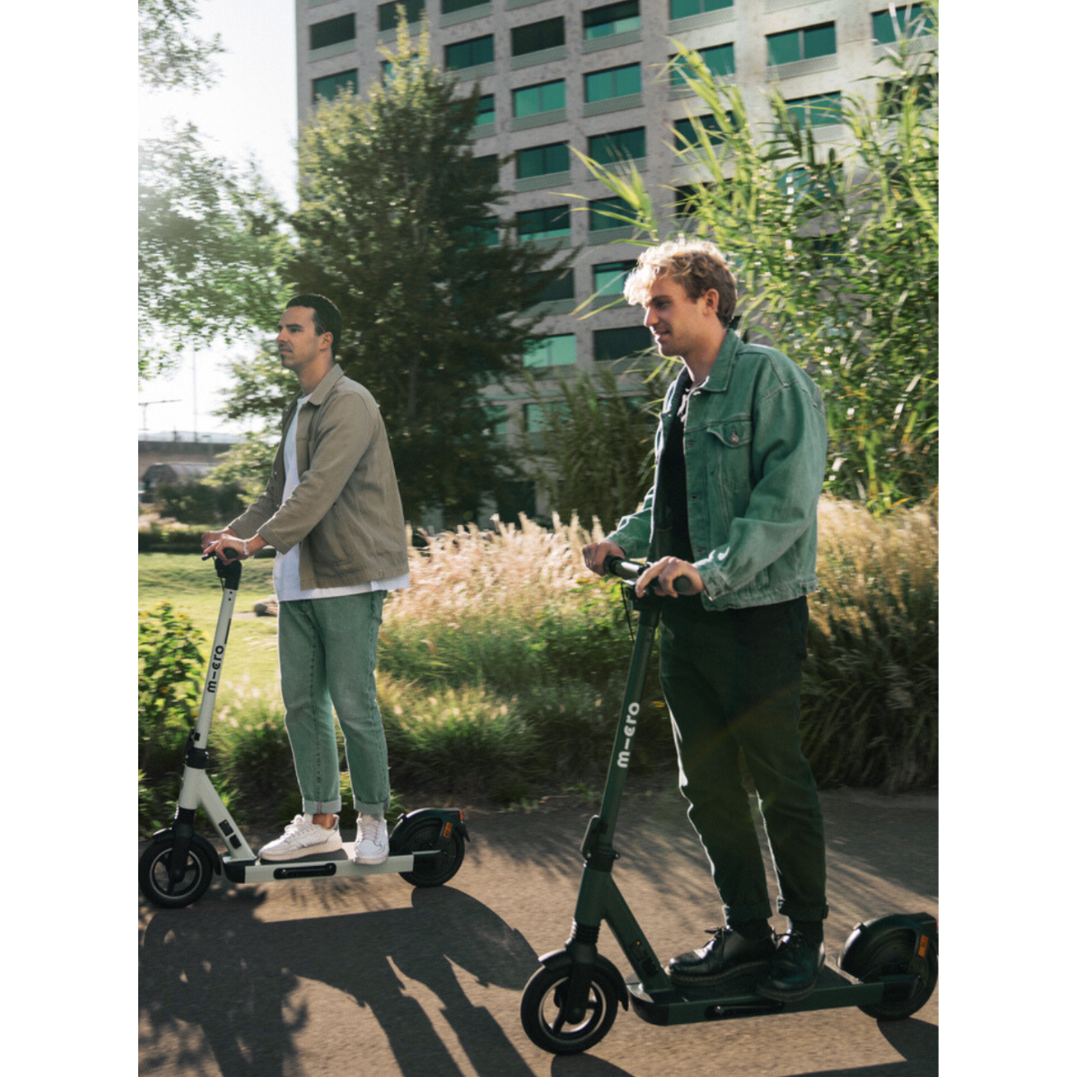 Zwei Männer fahren E-Scooter in einem Park mit einem Gebäude im Hintergrund.