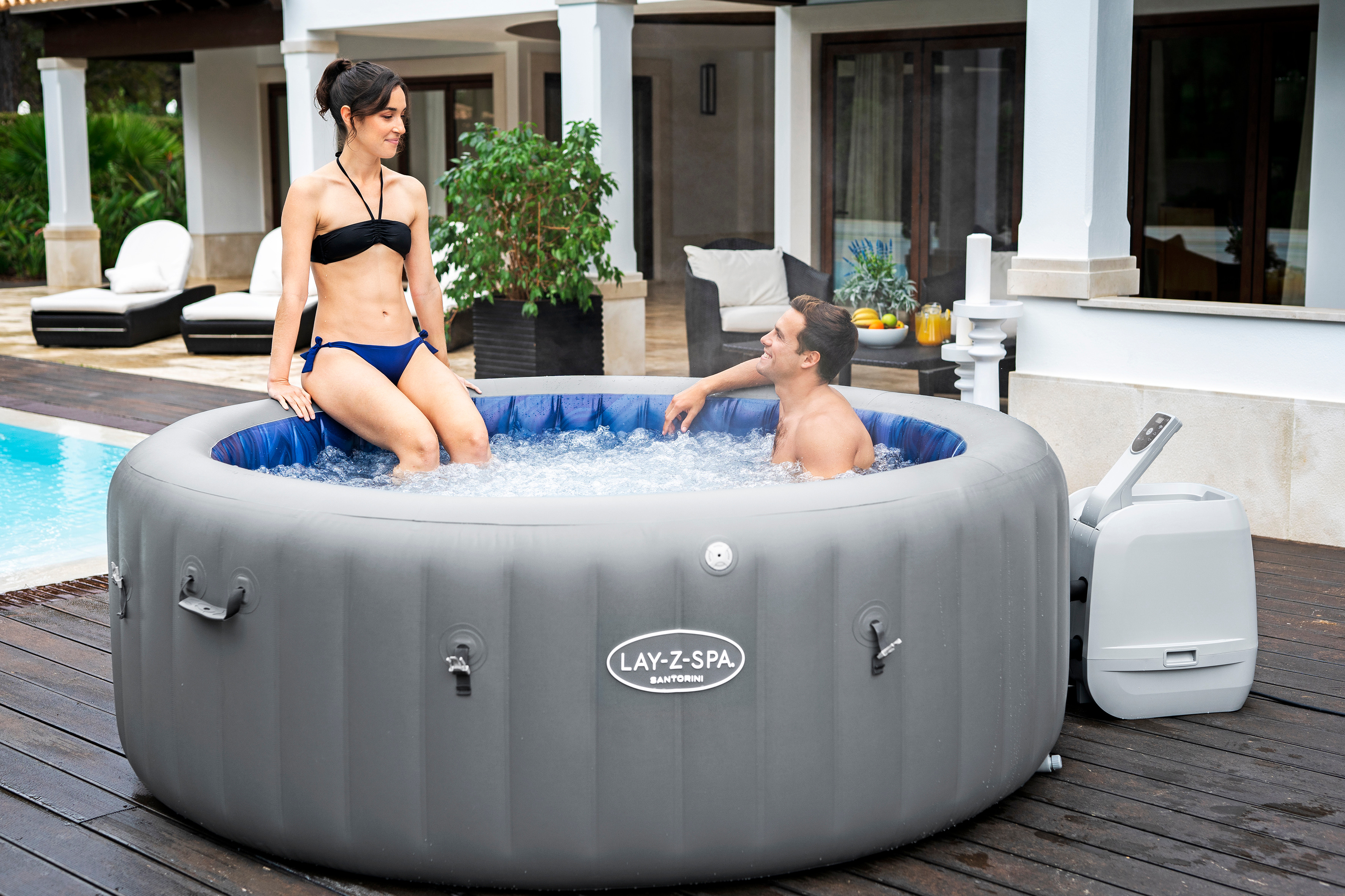 Ein Paar entspannt in einem aufblasbaren Whirlpool auf einer Holzterrasse neben einem Pool.