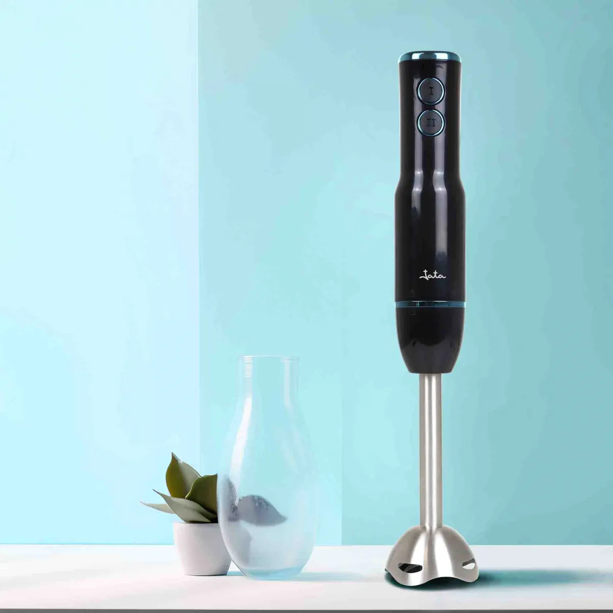 Czarny blender ręczny z srebrną łodygą obok szklanego wazonu i rośliny doniczkowej.