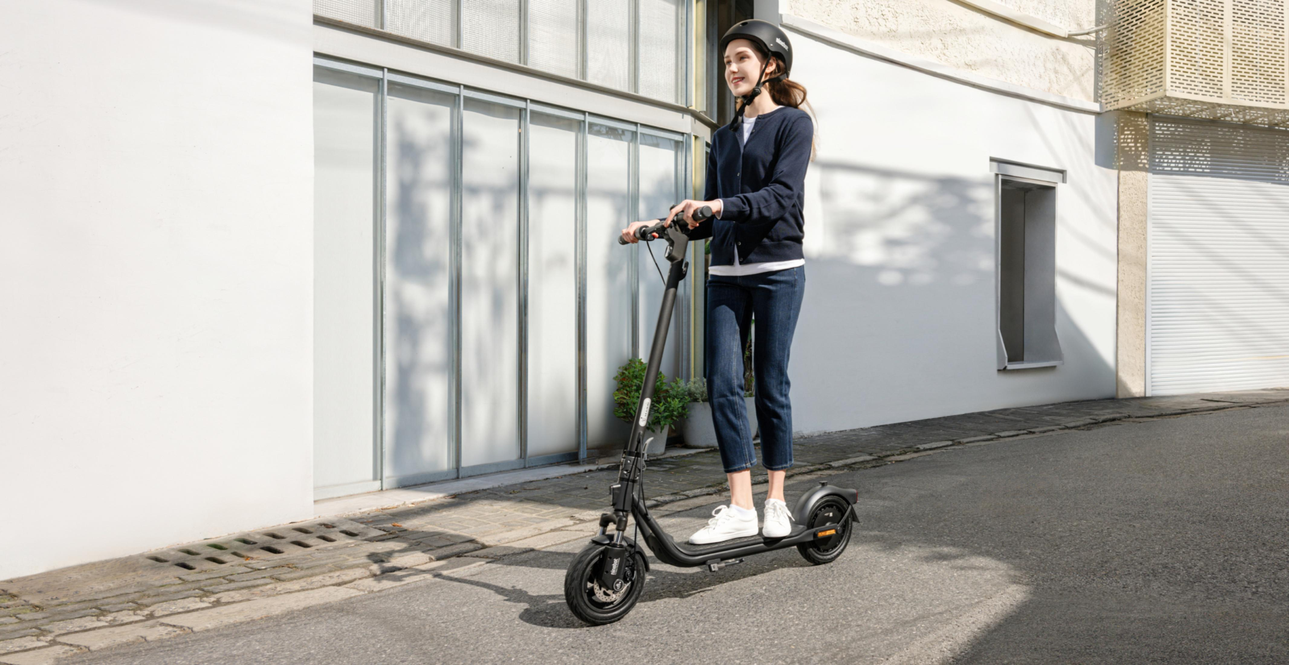 Eine Frau mit Helm fährt einen schwarzen Elektroroller auf einer Straße.