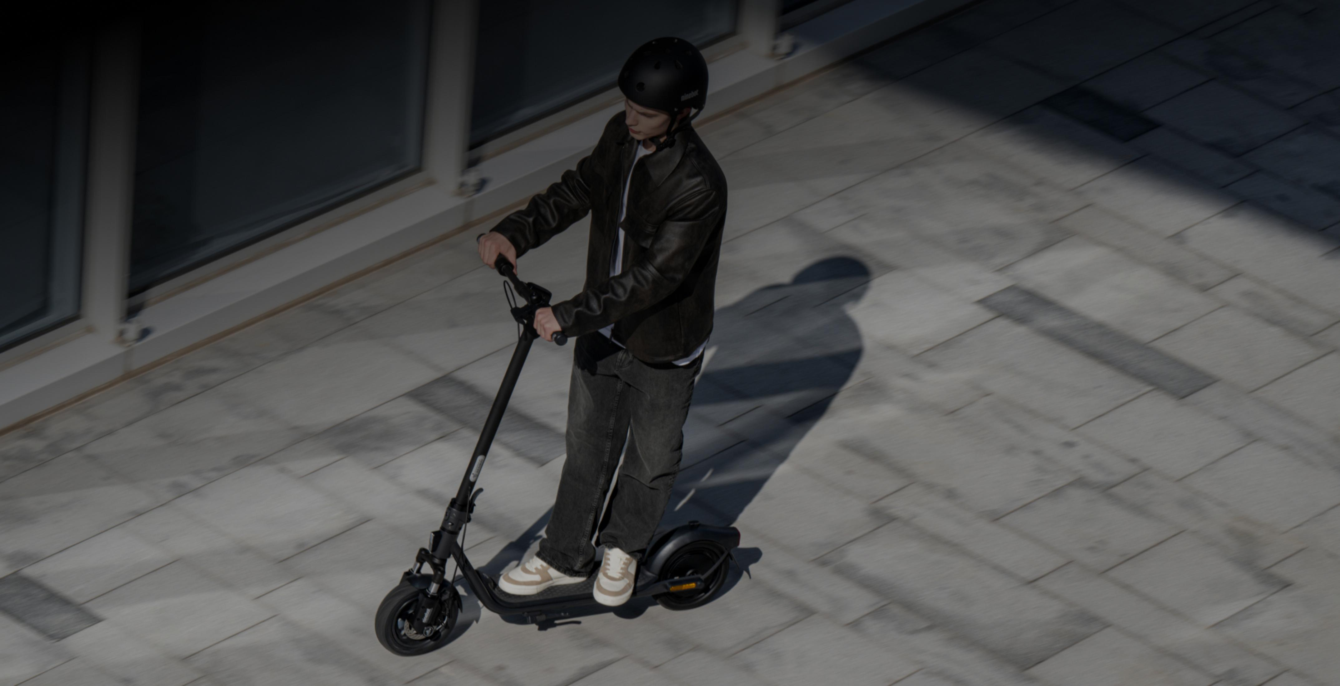Eine Person mit schwarzem Helm und Lederjacke fährt einen Elektroroller auf einem gepflasterten Weg.