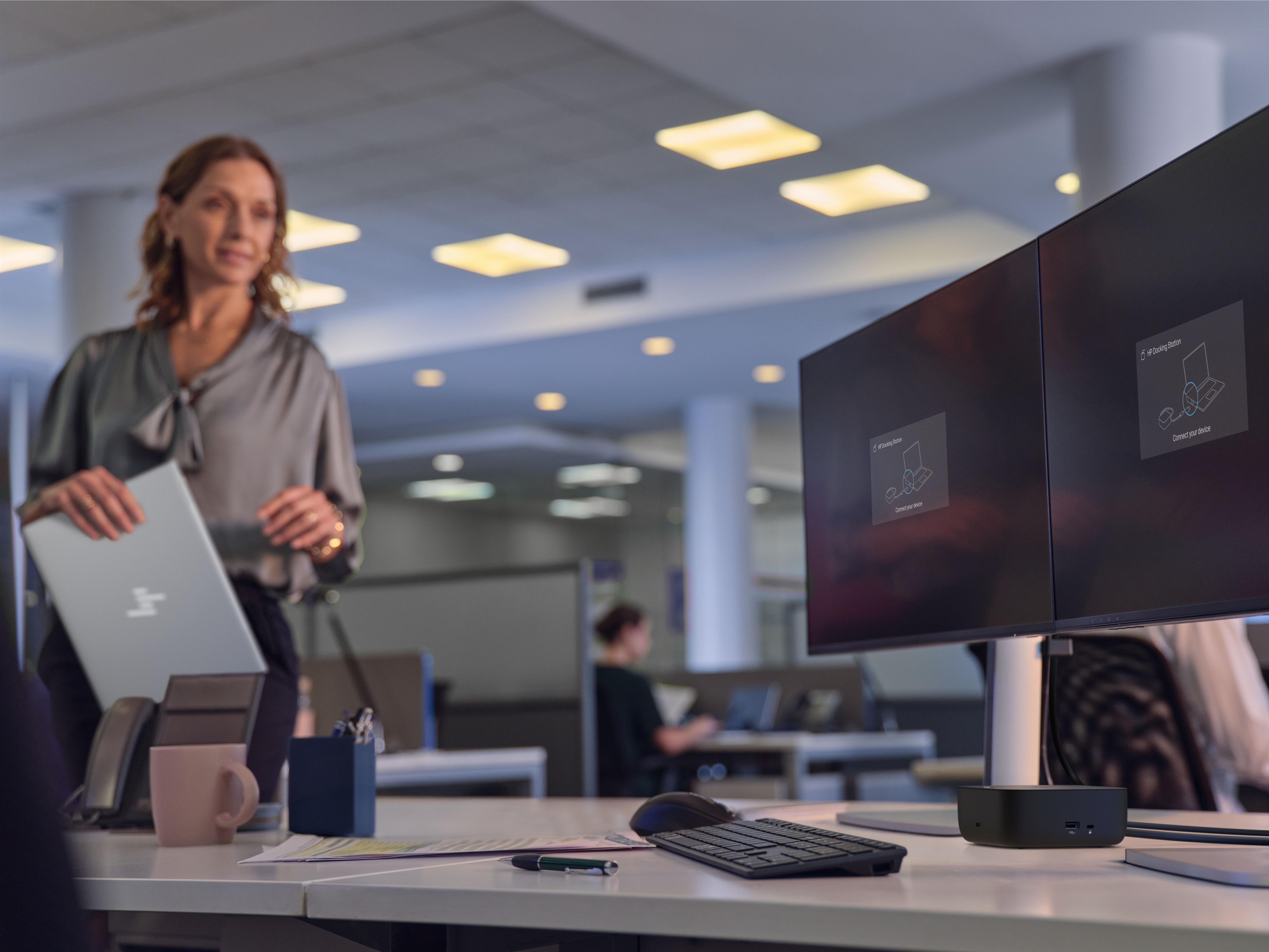 Eine Frau im Büro hält einen Laptop. Im Vordergrund sind Monitore und eine Dockingstation.