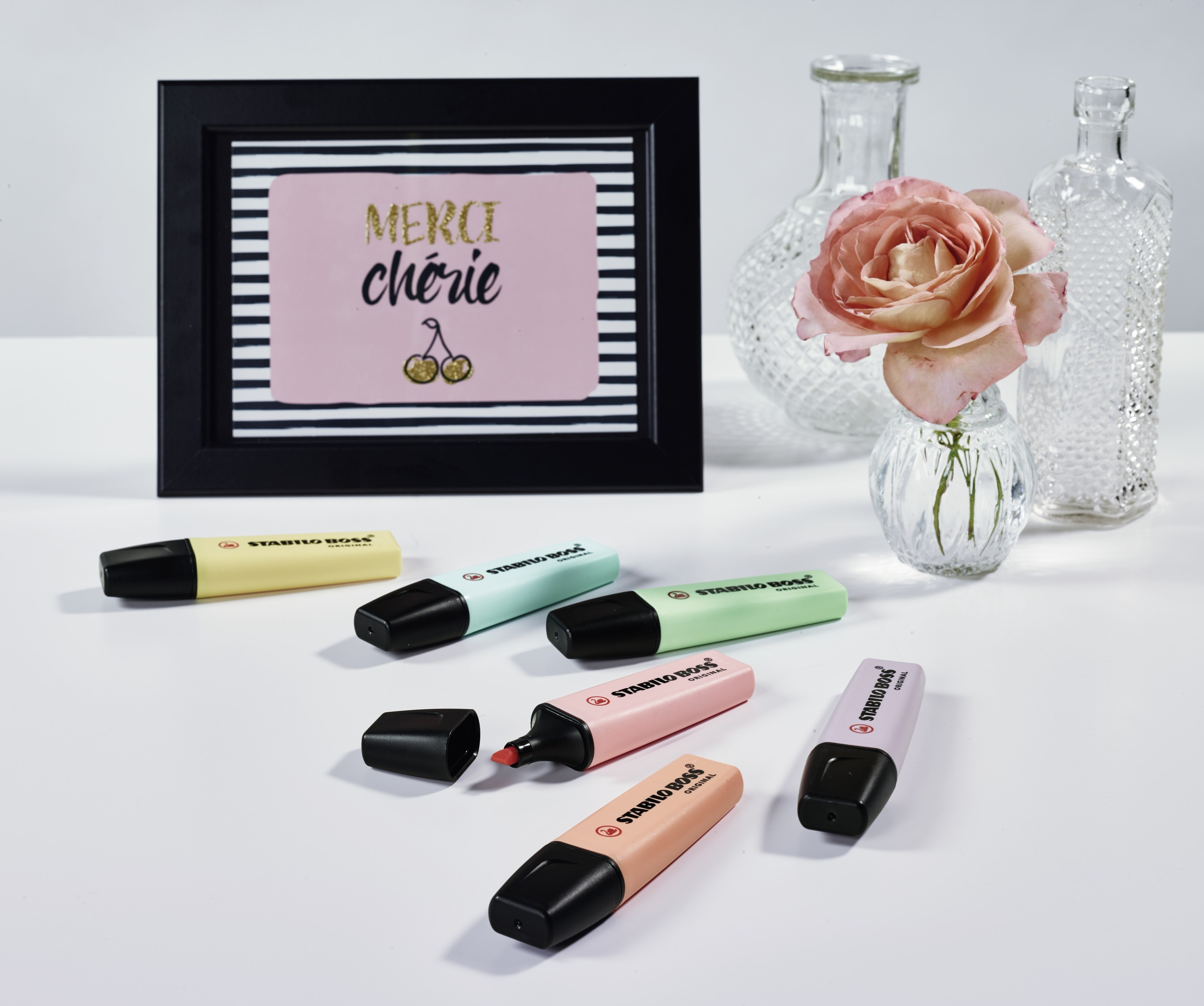Gerahmtes Bild mit Text, eine Rose in einer Vase und bunte Textmarker auf weißem Tisch.