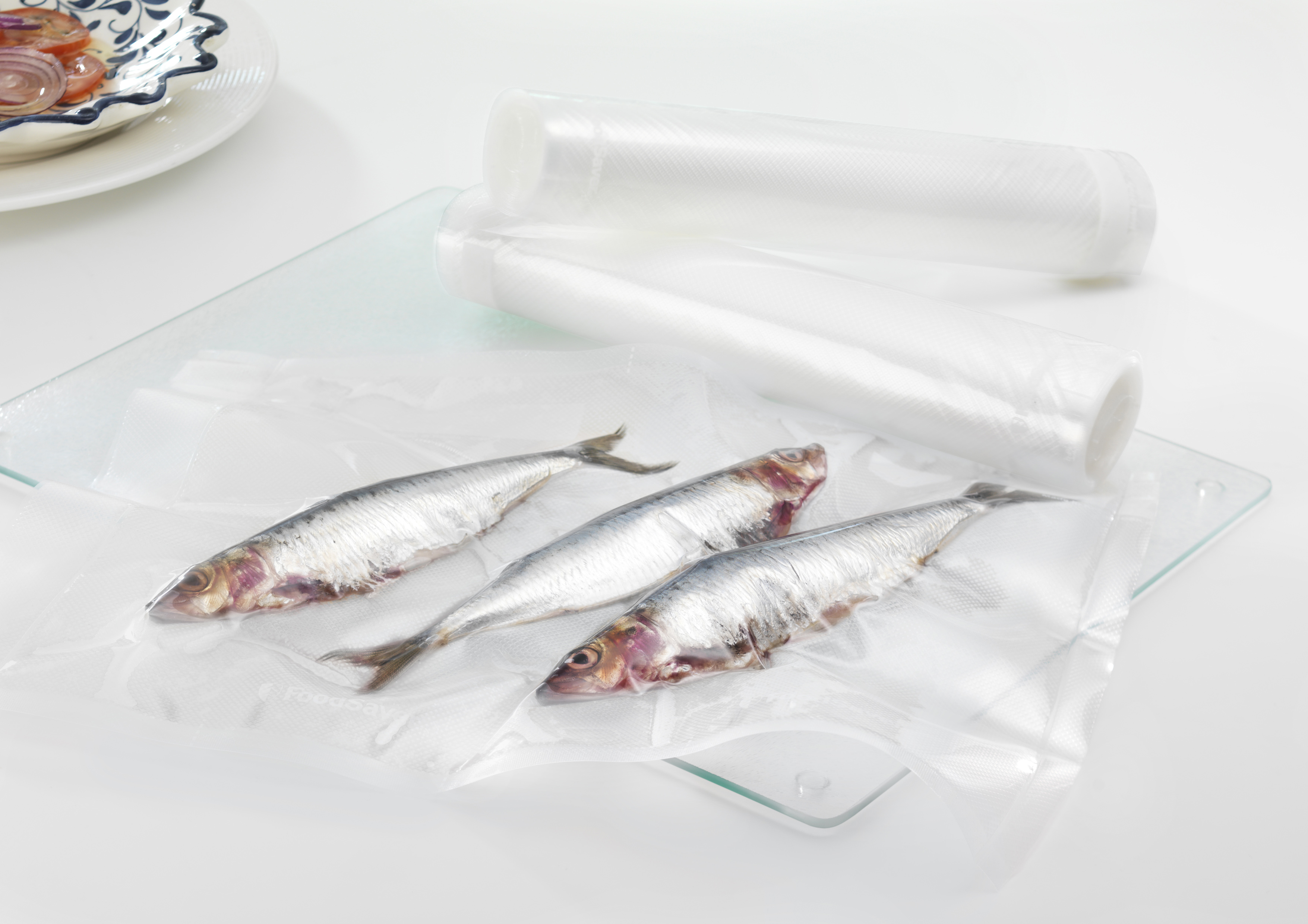 Drei rohe Heringe in einem Vakuumbeutel auf einer Glasplatte, neben Rollen mit Vakuumbeutelmaterial.