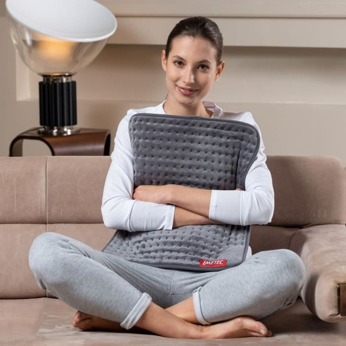 Mujer sentada en un sofá, sosteniendo una almohadilla térmica gris.