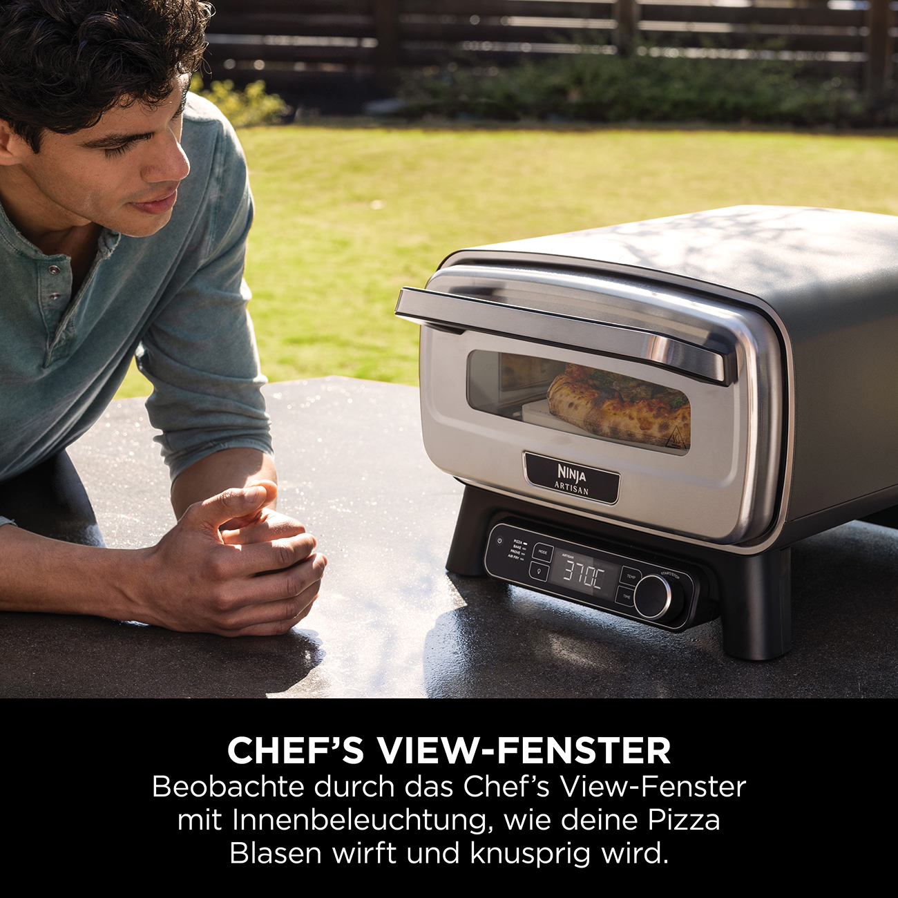 Man looks at pizza oven with pizza inside. Grass and fence in background.
