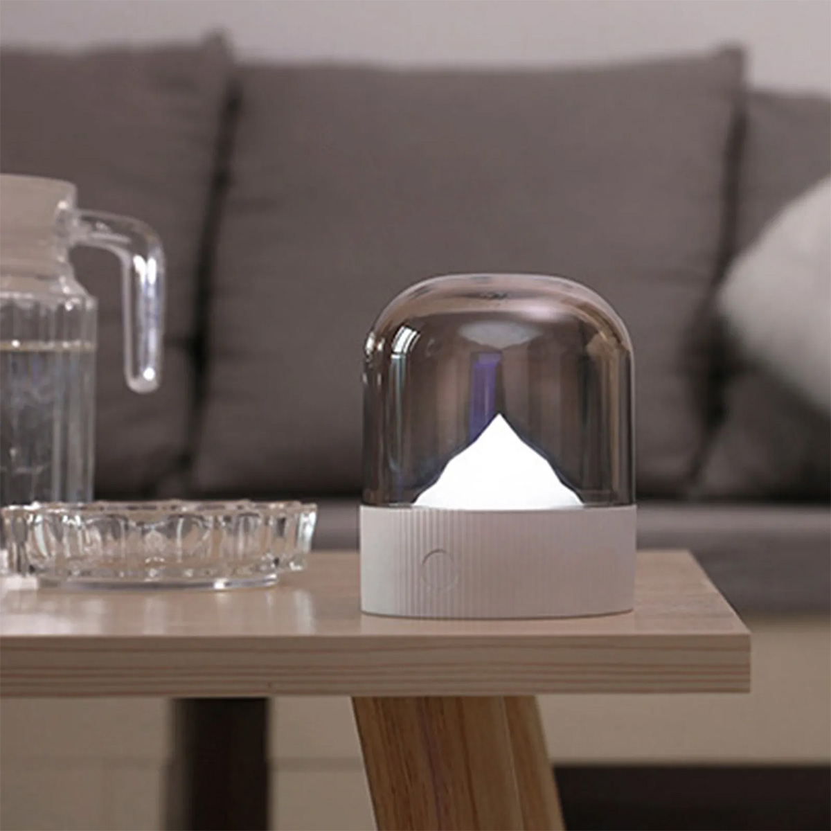 Eine weiße Lampe mit Bergform unter einer durchsichtigen Kuppel auf einem Holztisch.