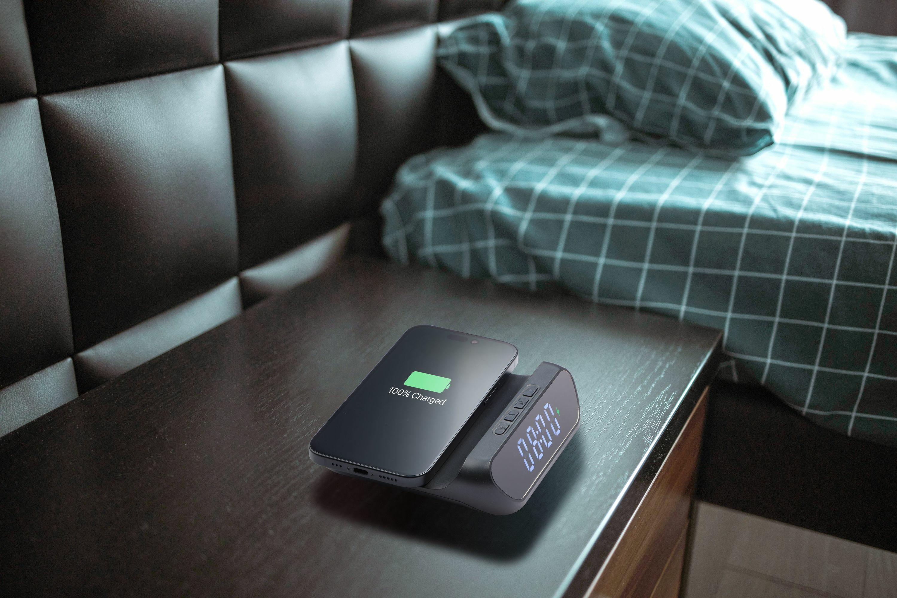 Un smartphone con 100% de carga en una mesita de noche junto a una cama.