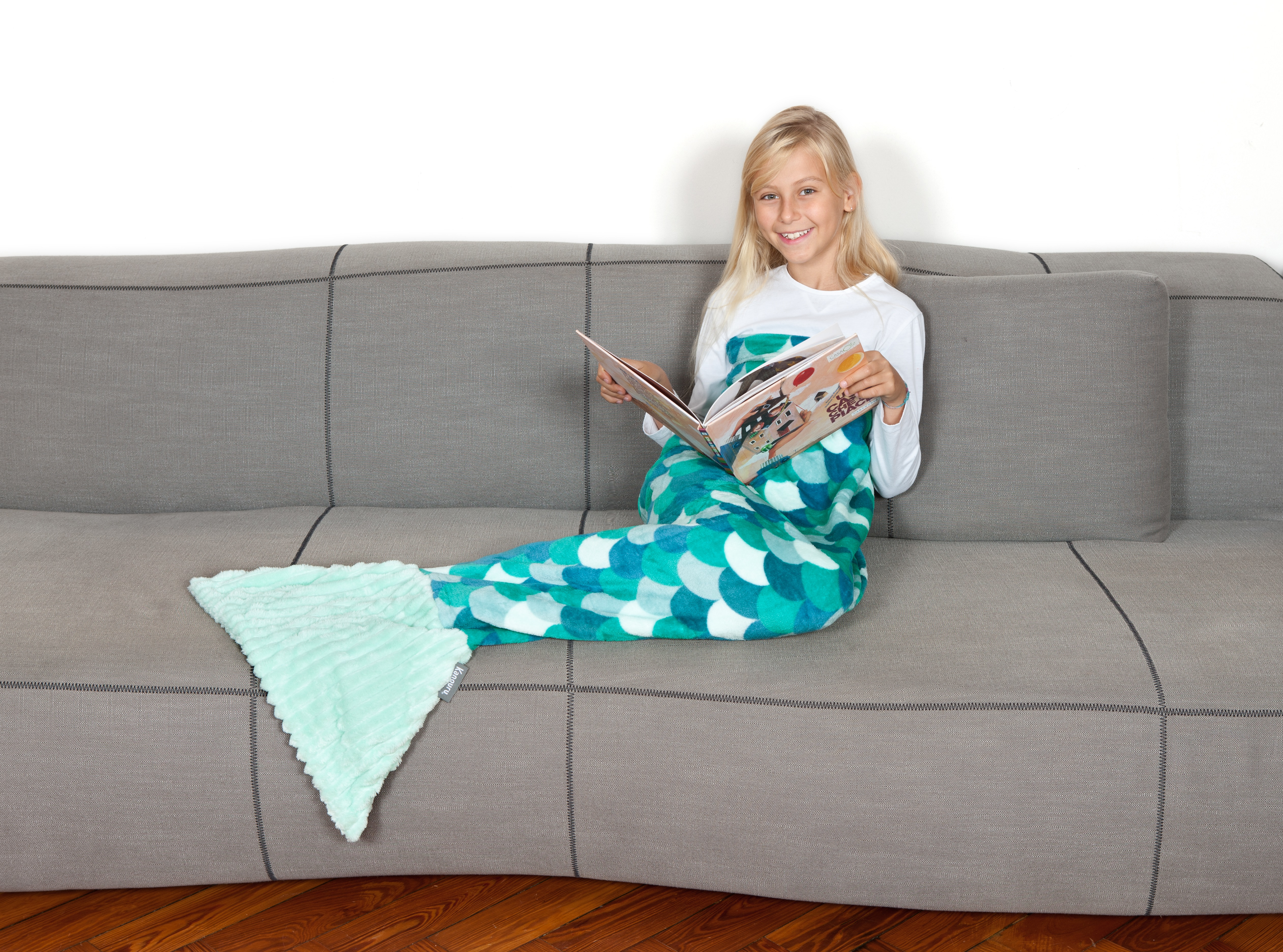 Una ragazza con una coperta a coda di sirena legge un libro sul divano.